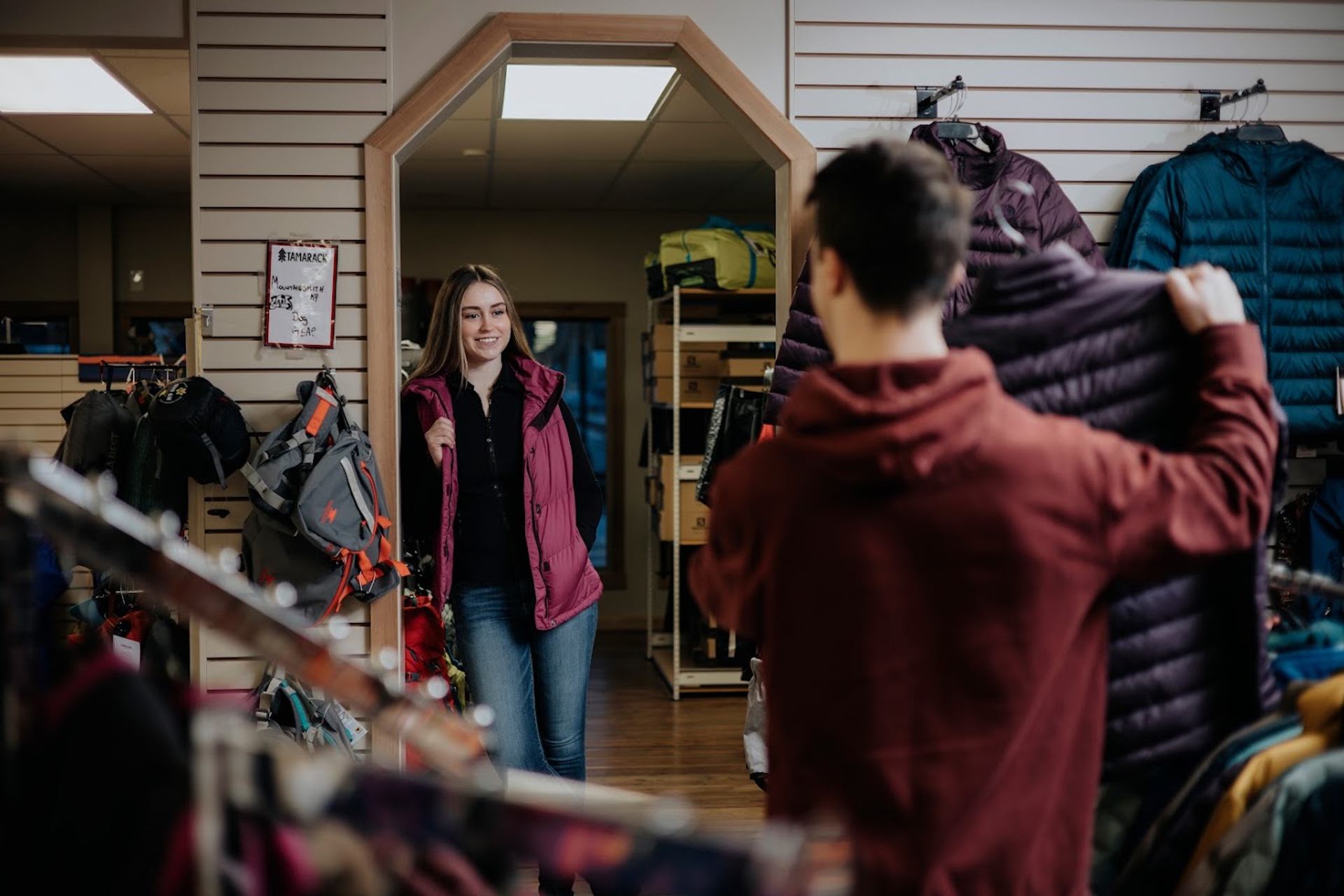 Outdoor gear store interior with shoppers browsing jackets and backpacks on wall displays.