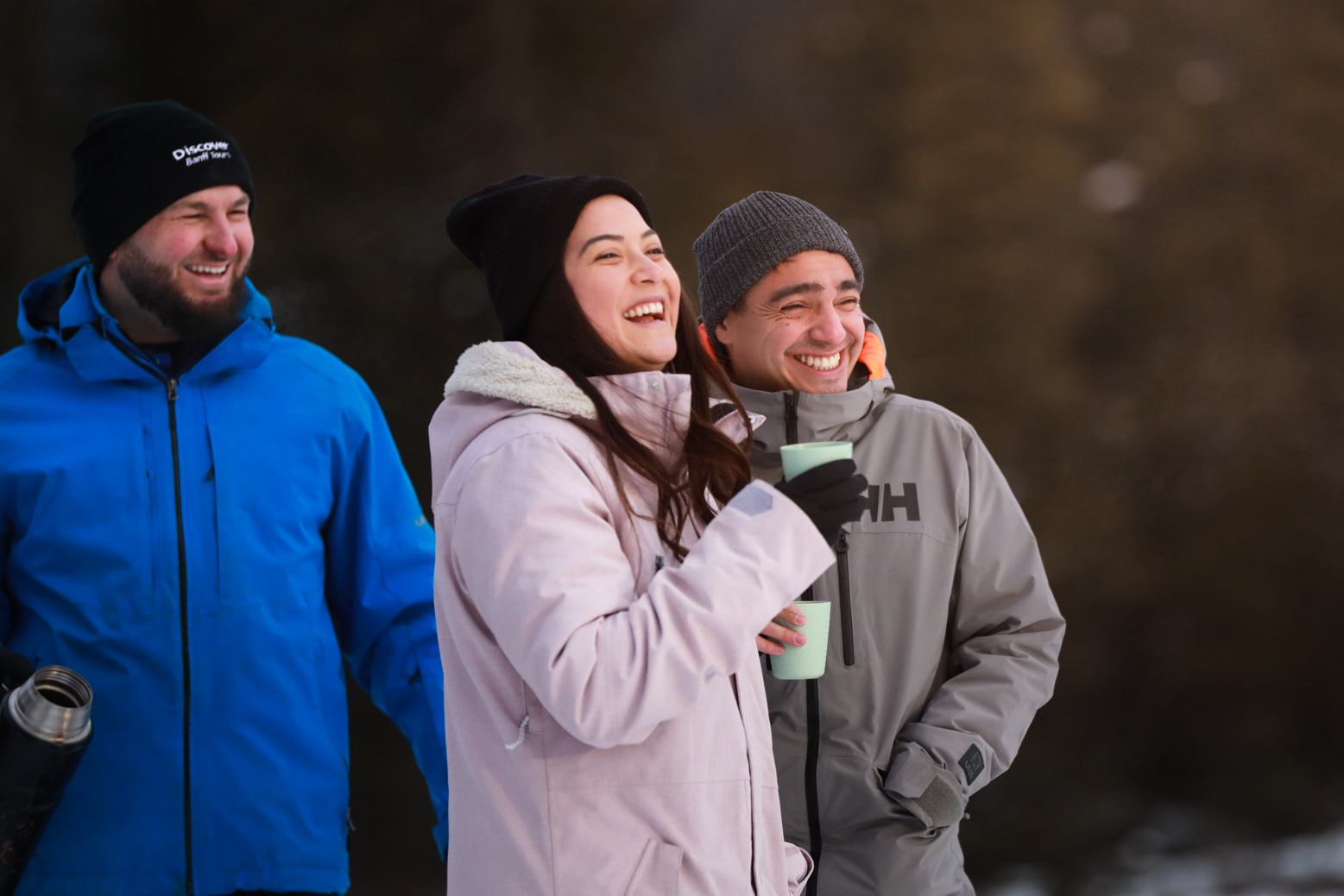Three people in winter jackets standing outdoors with snow on the ground and trees in the background.