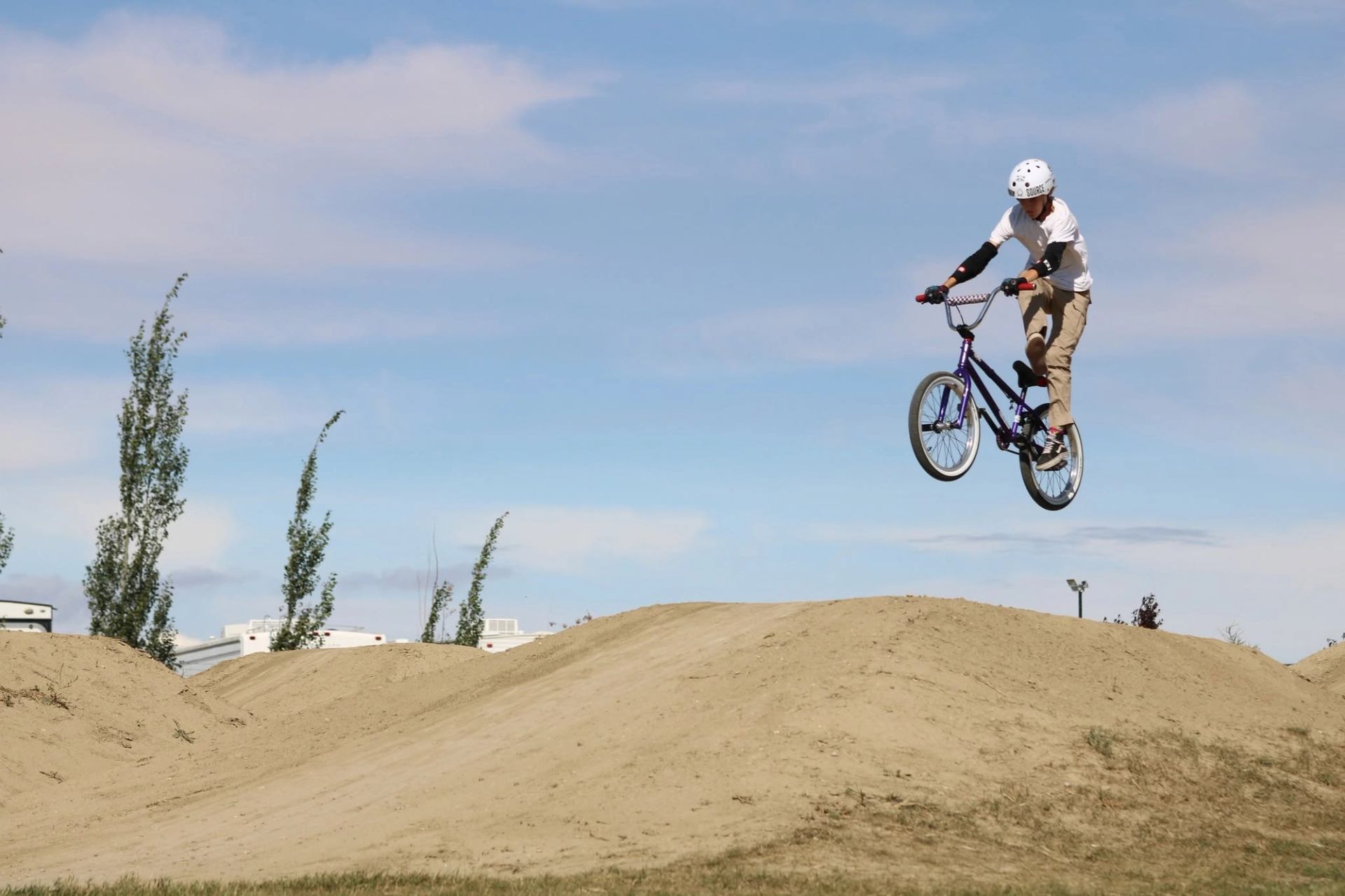 BMX rider wearing helmet jumps off dirt ramp against blue sky at bike park.