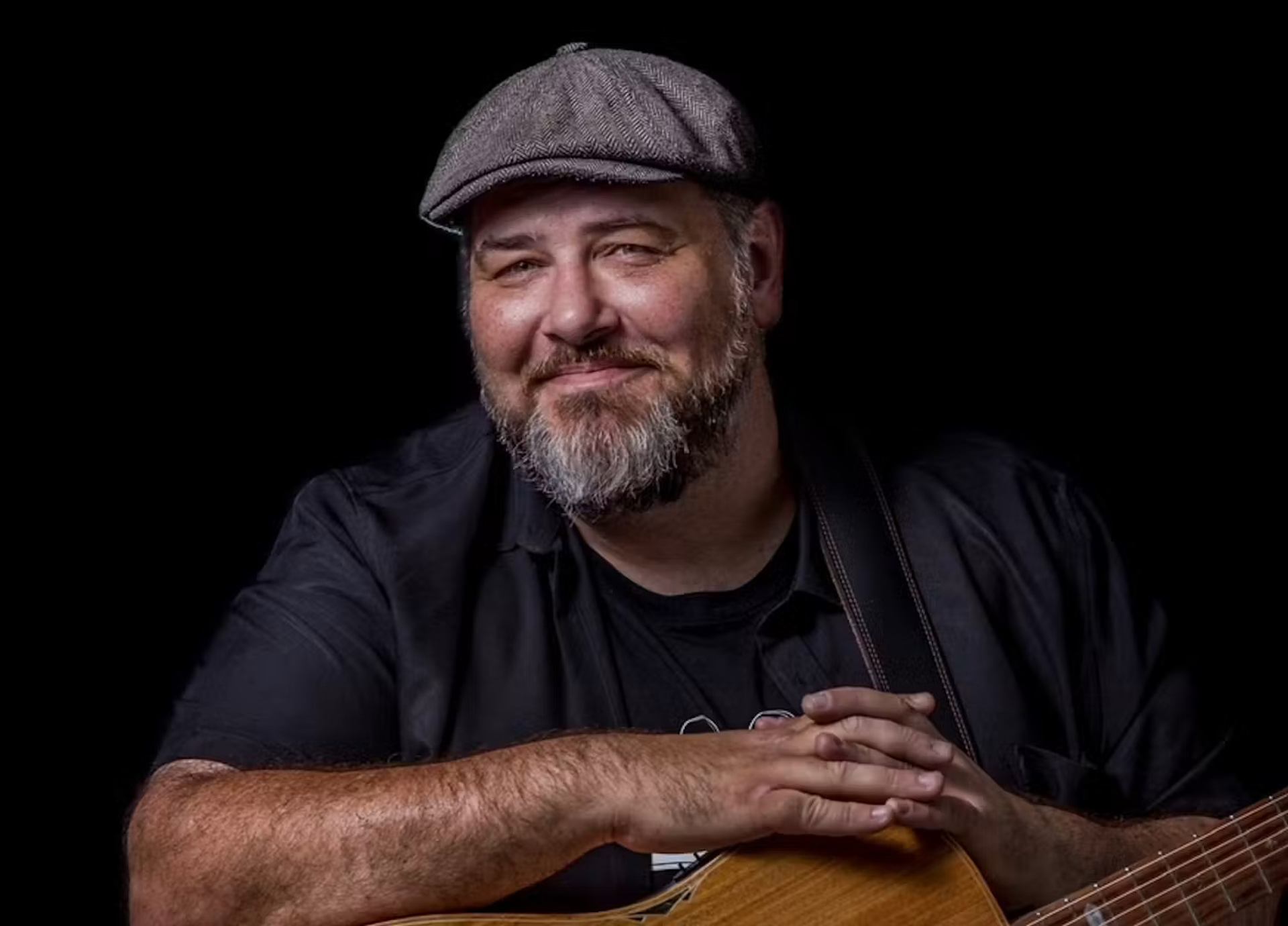 Bearded man in flat cap rests hands on acoustic guitar against black background.