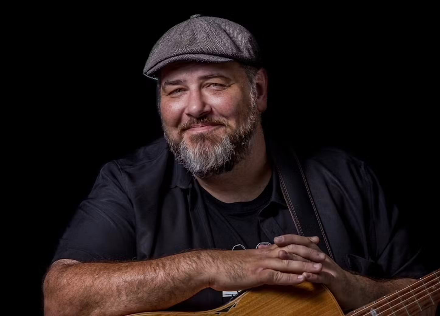 Bearded man in flat cap rests hands on acoustic guitar against black background.