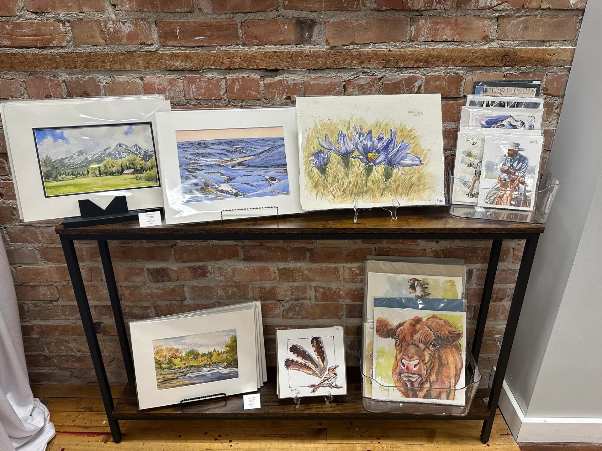 Framed prints and loose artworks displayed on shelves against a brick wall.