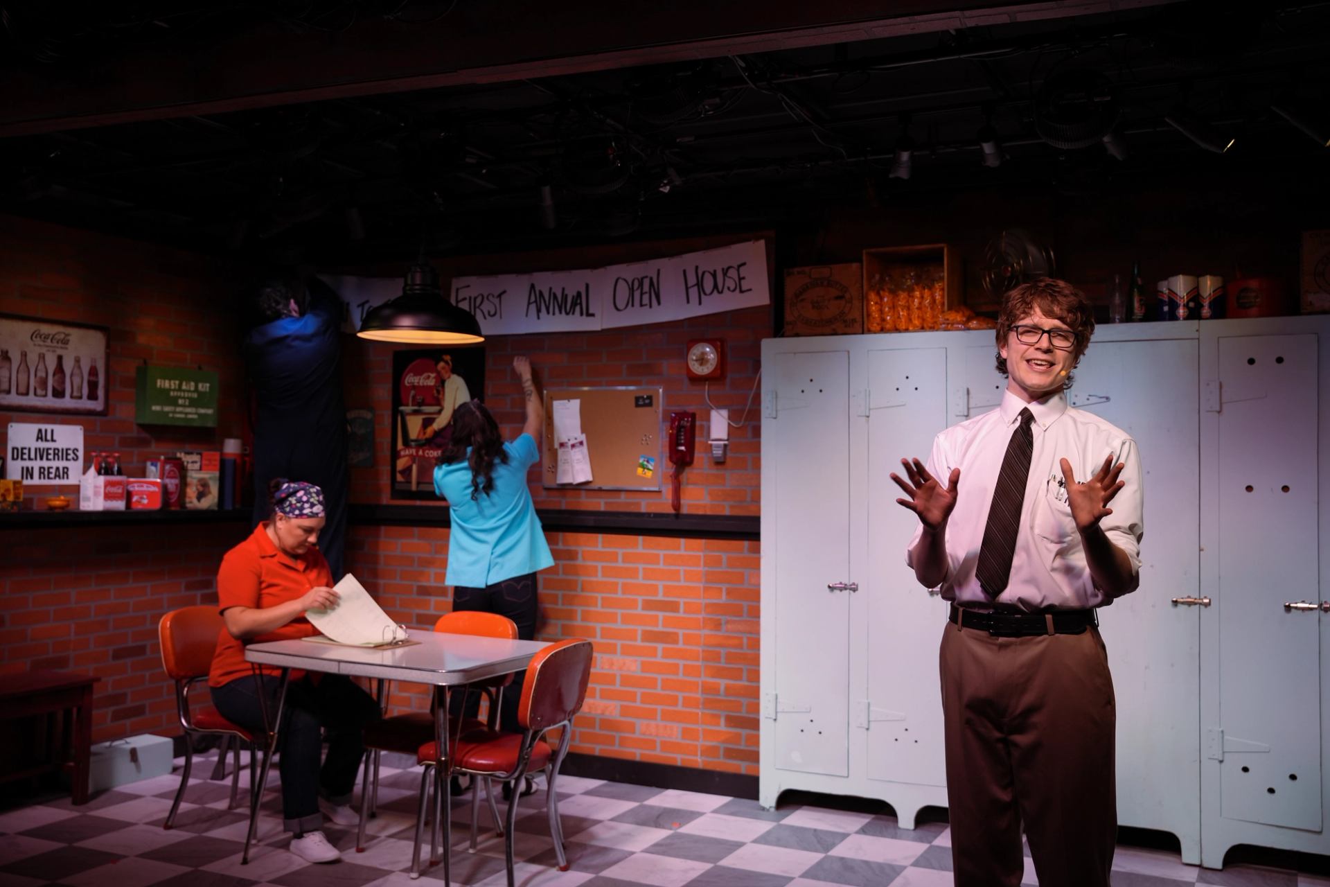 Stage scene in staff room set, workers decorate while man in tie speaks.