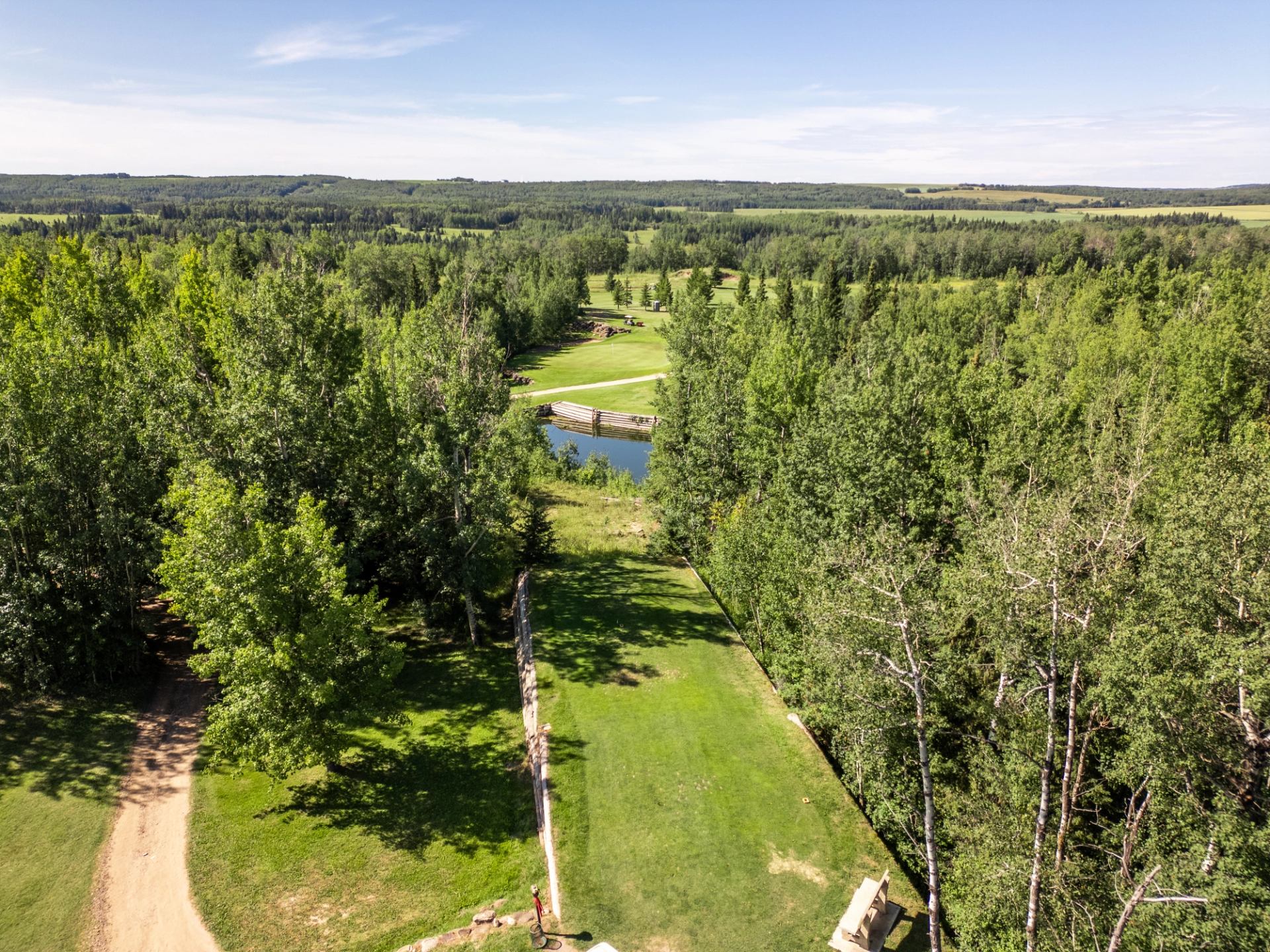 Aerial view of a long green range bordered by dense forest, with a small pond and distant fields under a clear sky.