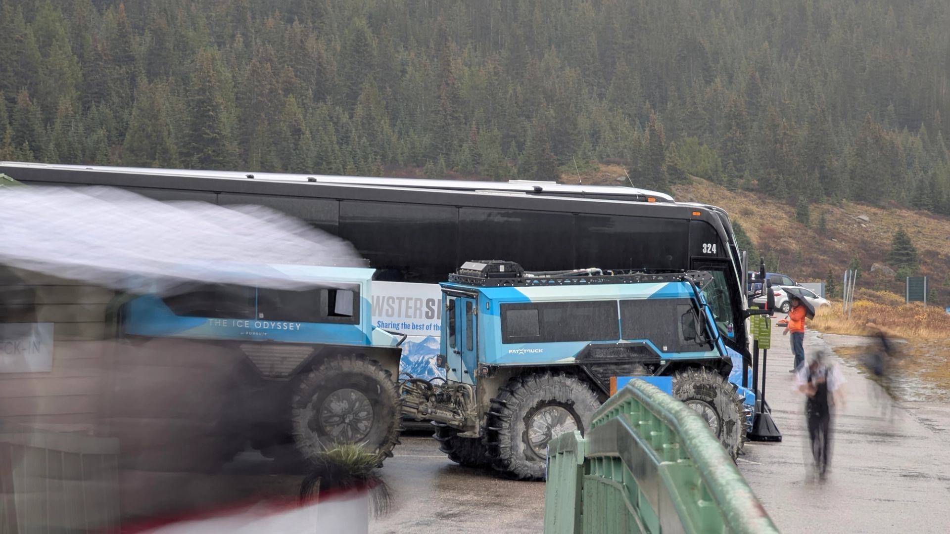 Blue off-road vehicle and tour bus parked near forested mountains on a cloudy day.