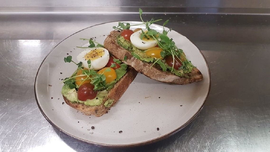 Two slices of toast topped with avocado, cherry tomatoes, sliced boiled egg, and microgreens on a white plate, creating a fresh, vibrant dish.