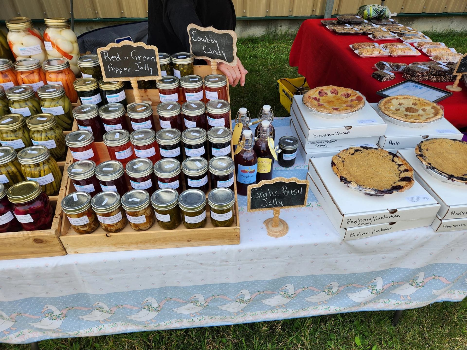 Market vendors display homemade pies, preserves, jellies, pickles, and baked goods on outdoor tables.