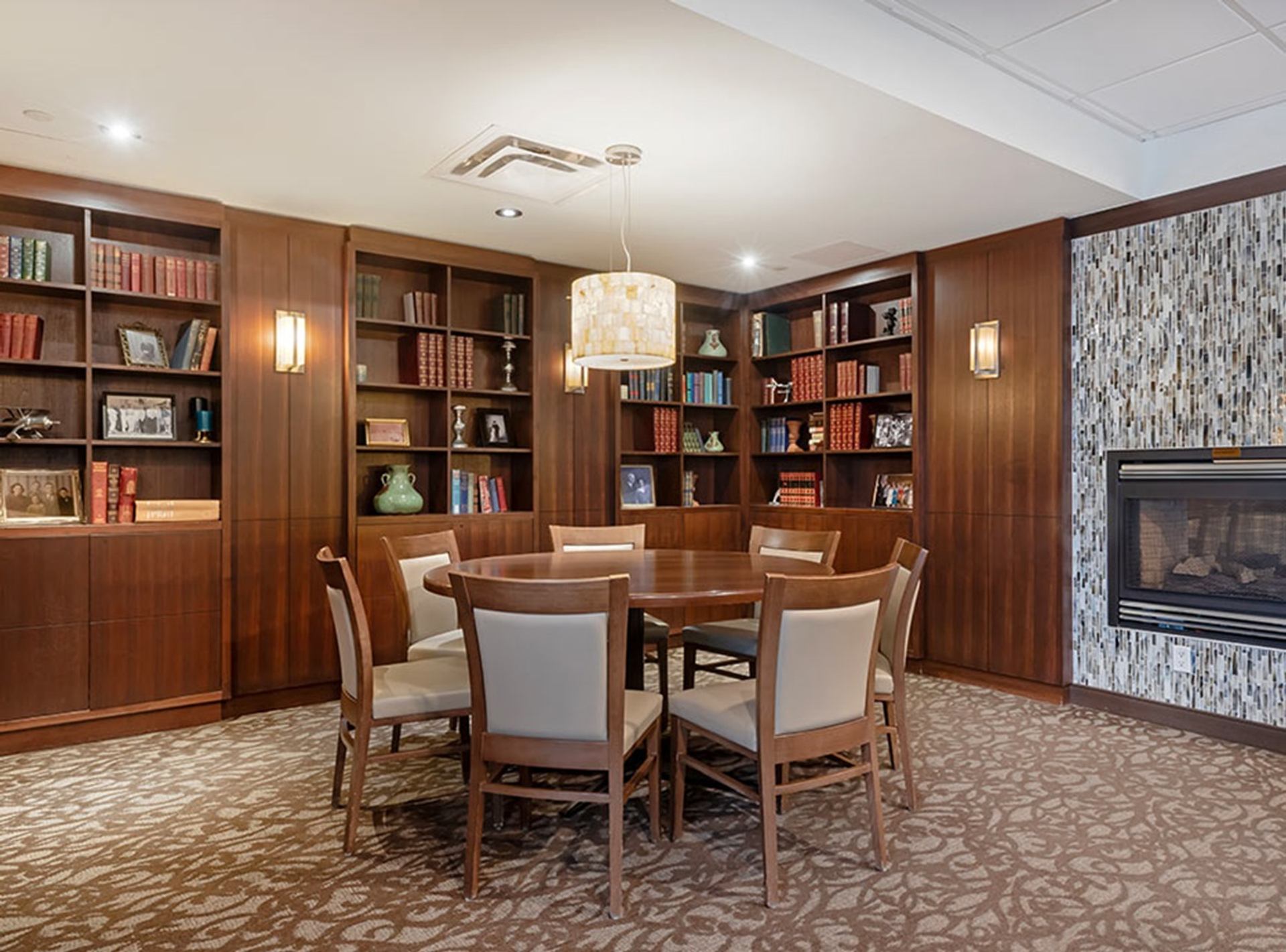 Library-style lounge with round wooden table, upholstered chairs, bookshelves and gas fireplace.