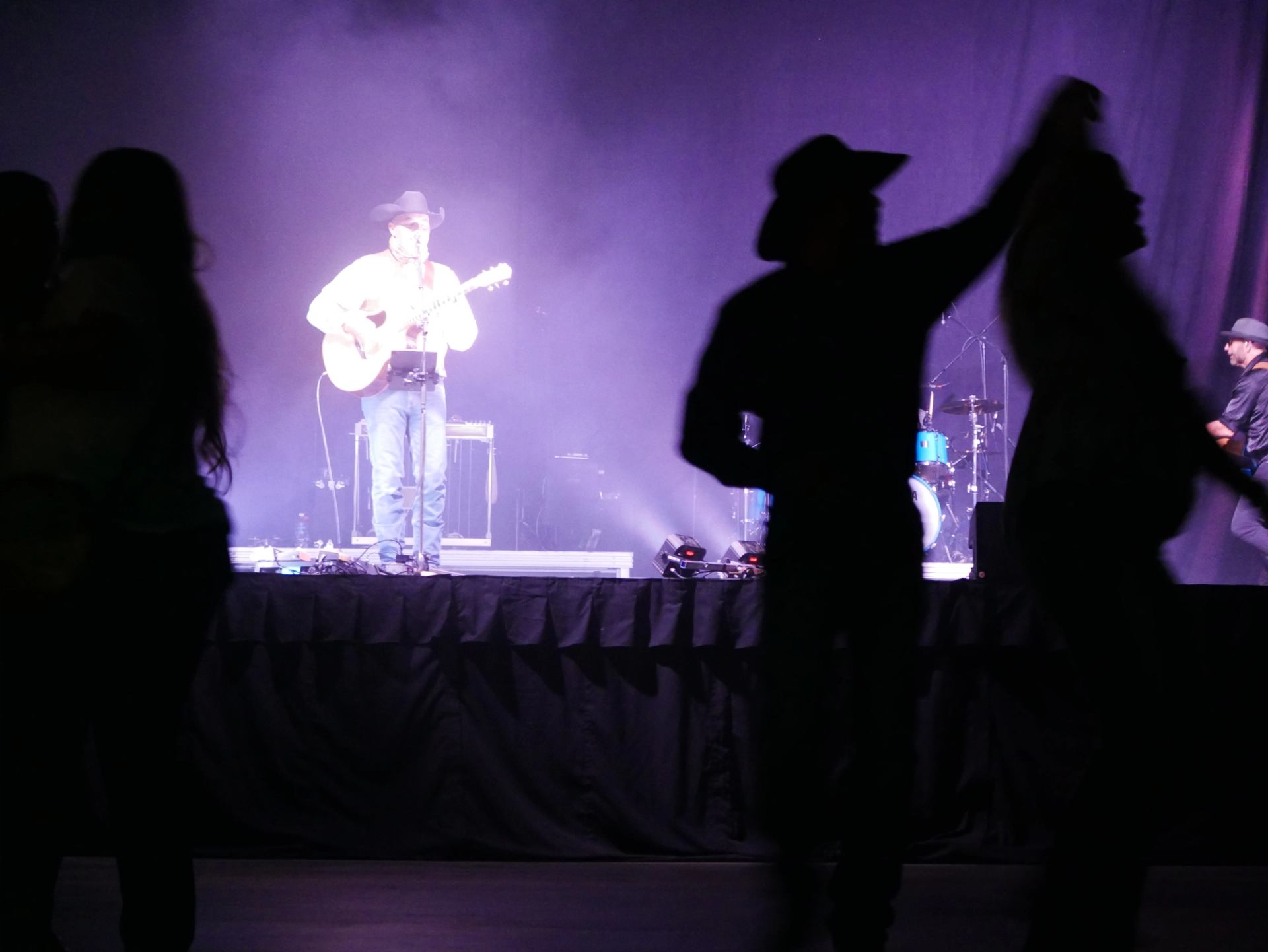 A country musician plays guitar on a purple-lit stage while silhouetted people dance in the foreground.