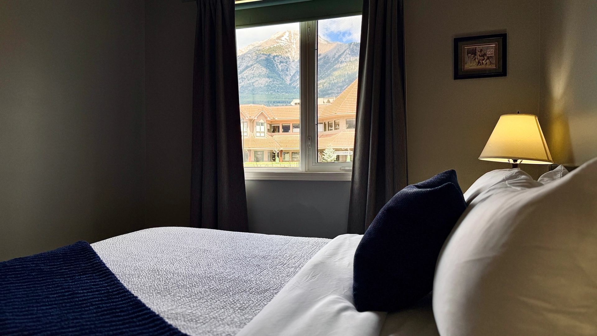 Bedroom with blue accents and a lit lamp overlooking buildings and snow-capped mountains.