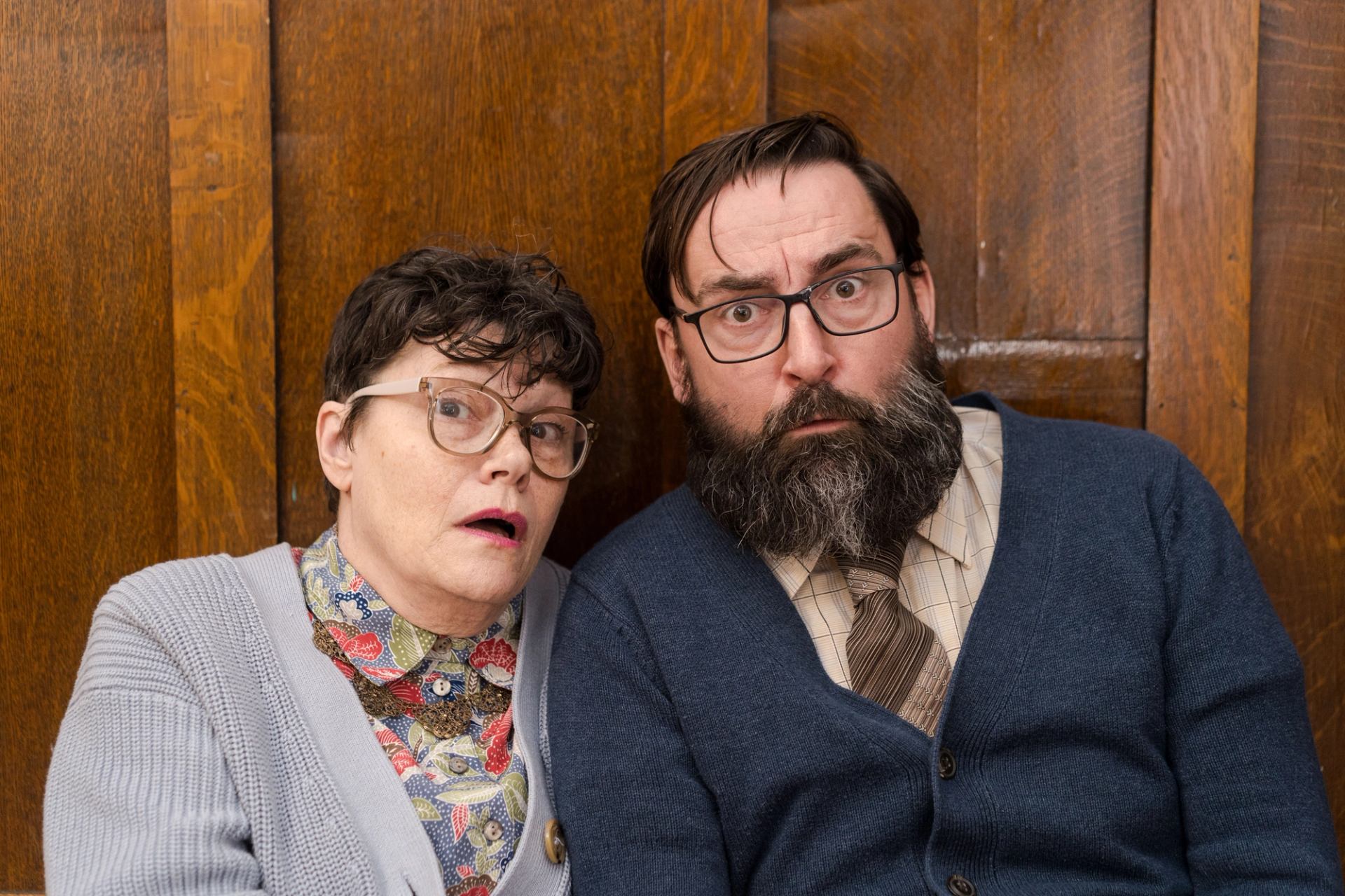 Two people seated closely against a wooden paneled wall, wearing vintage-style clothing.
