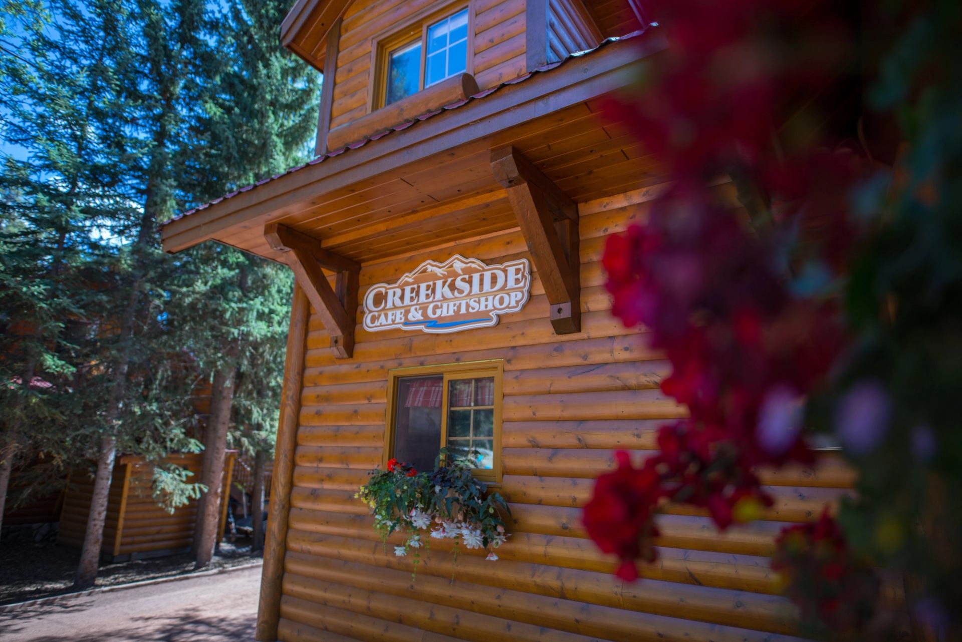 Creekside Cafe & Giftshop sign on log building, trees and blurred red flowers.