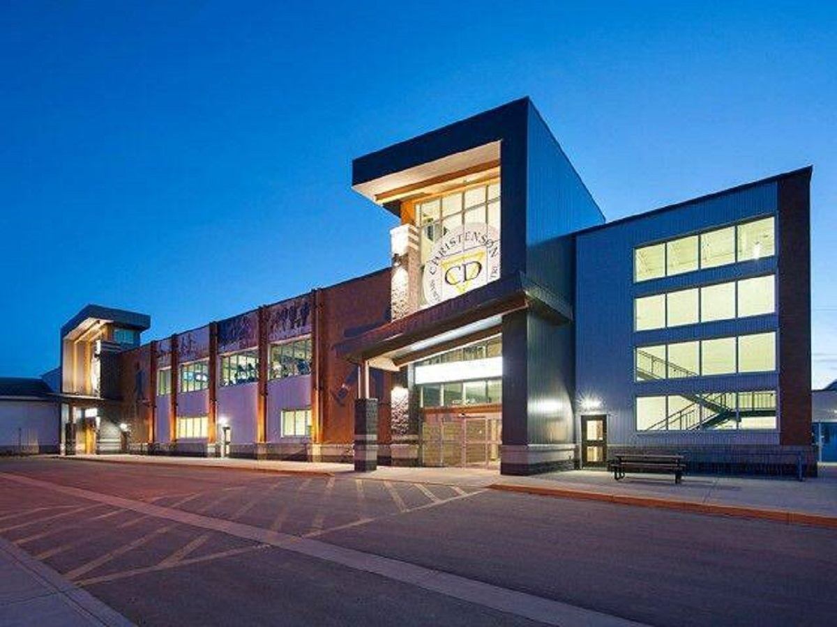 Modern school building with large lit windows and empty paved area at dusk.