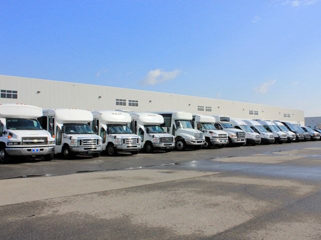 Row of white CST Transportations trucks parked outside warehouse under clear blue sky.
