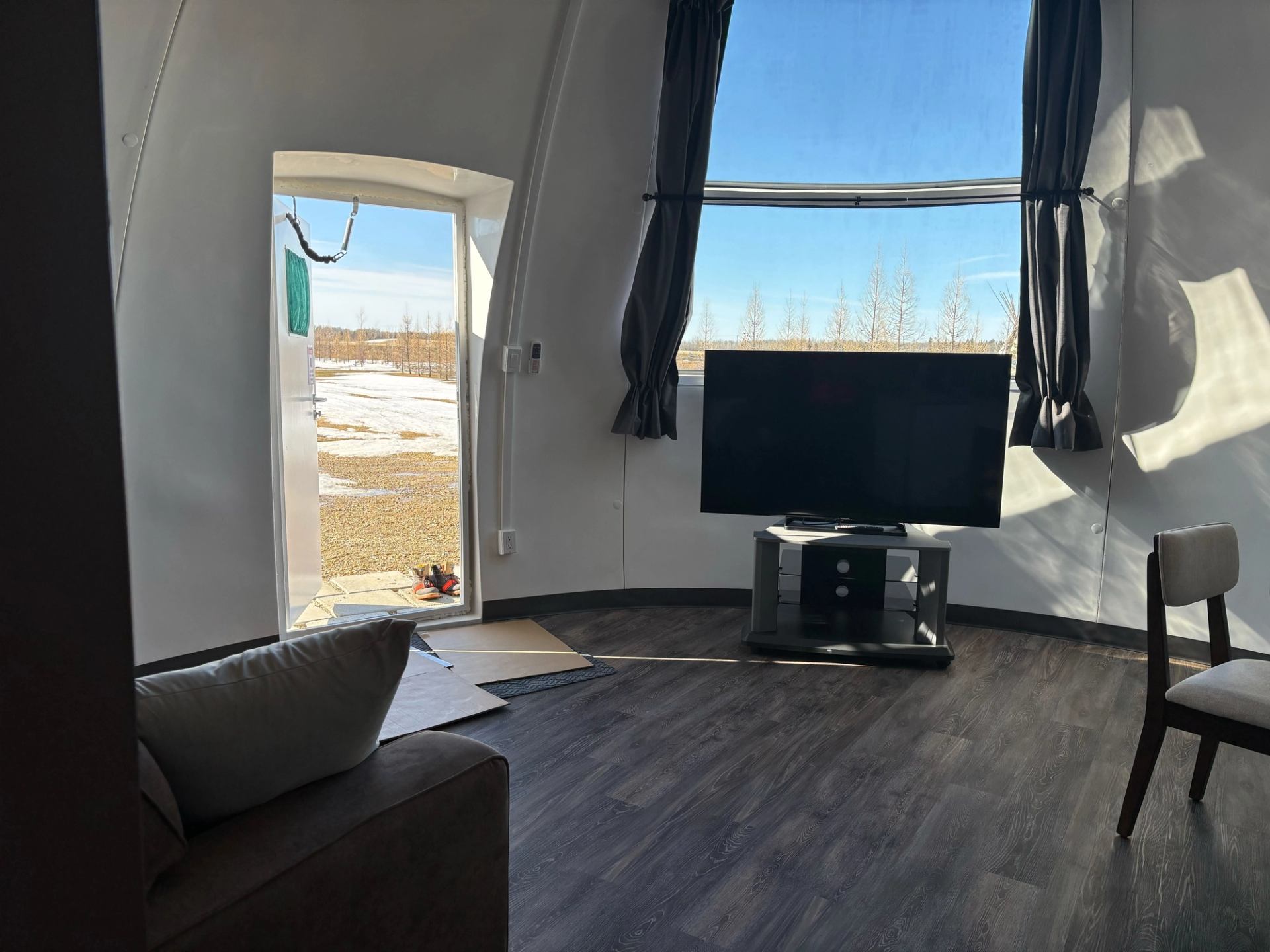 Dome interior with TV, window view, and cozy seating in snowy setting.