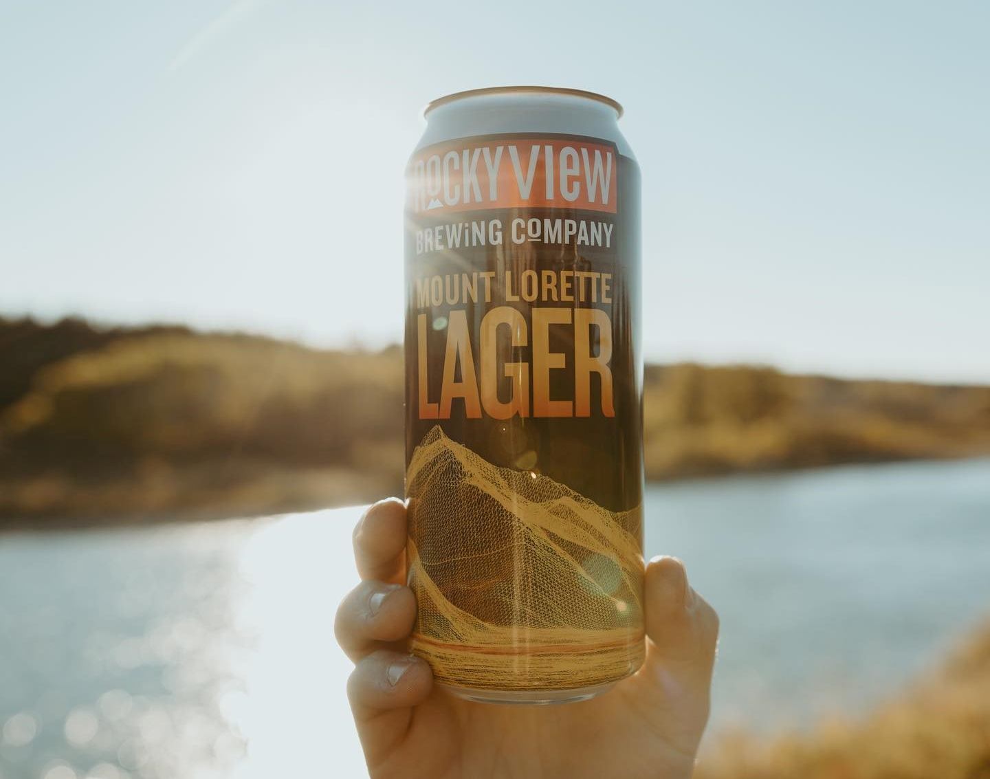 A can of Rocky View Brew held up with a lake and sunshine in the background.