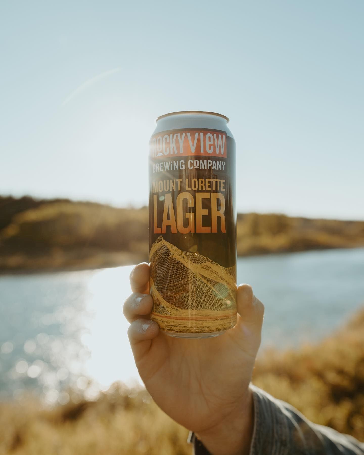 A can of Rocky View Brew held up with a lake and sunshine in the background.