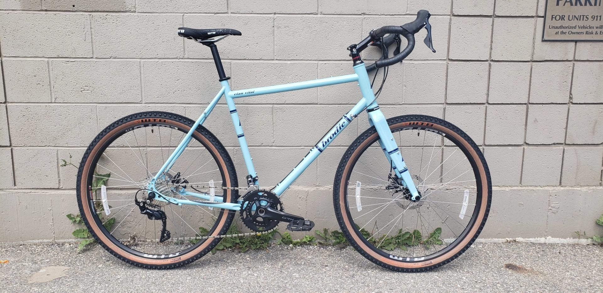 Light blue bike with brown rims by a beige brick wall and green plants.