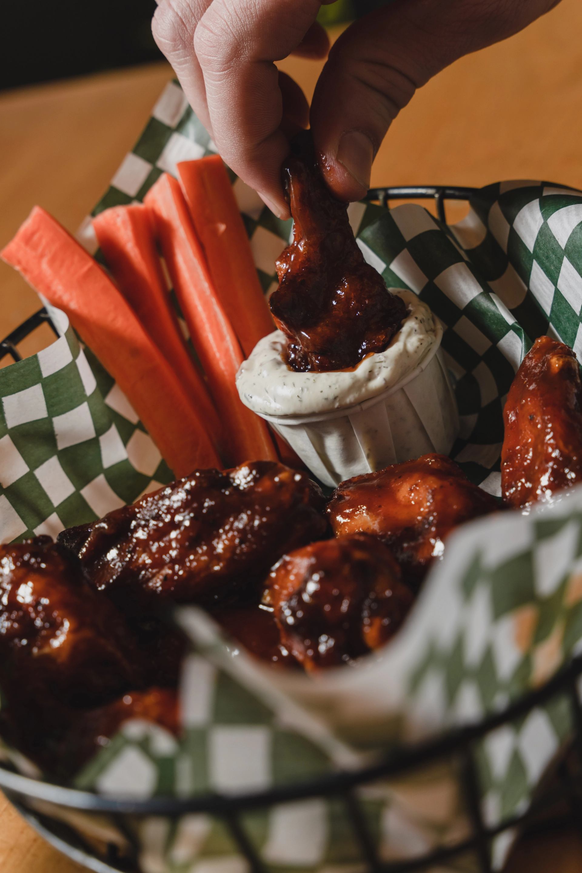 Hand dipping a saucy chicken wing into a white dip, served with carrot sticks in a basket.