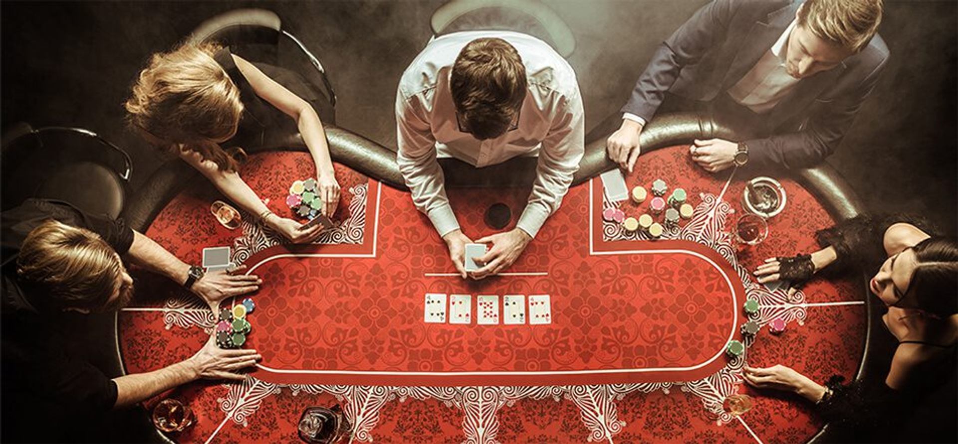 Five players are seated around a poker table featuring playing cards, chips, and a red felt surface.