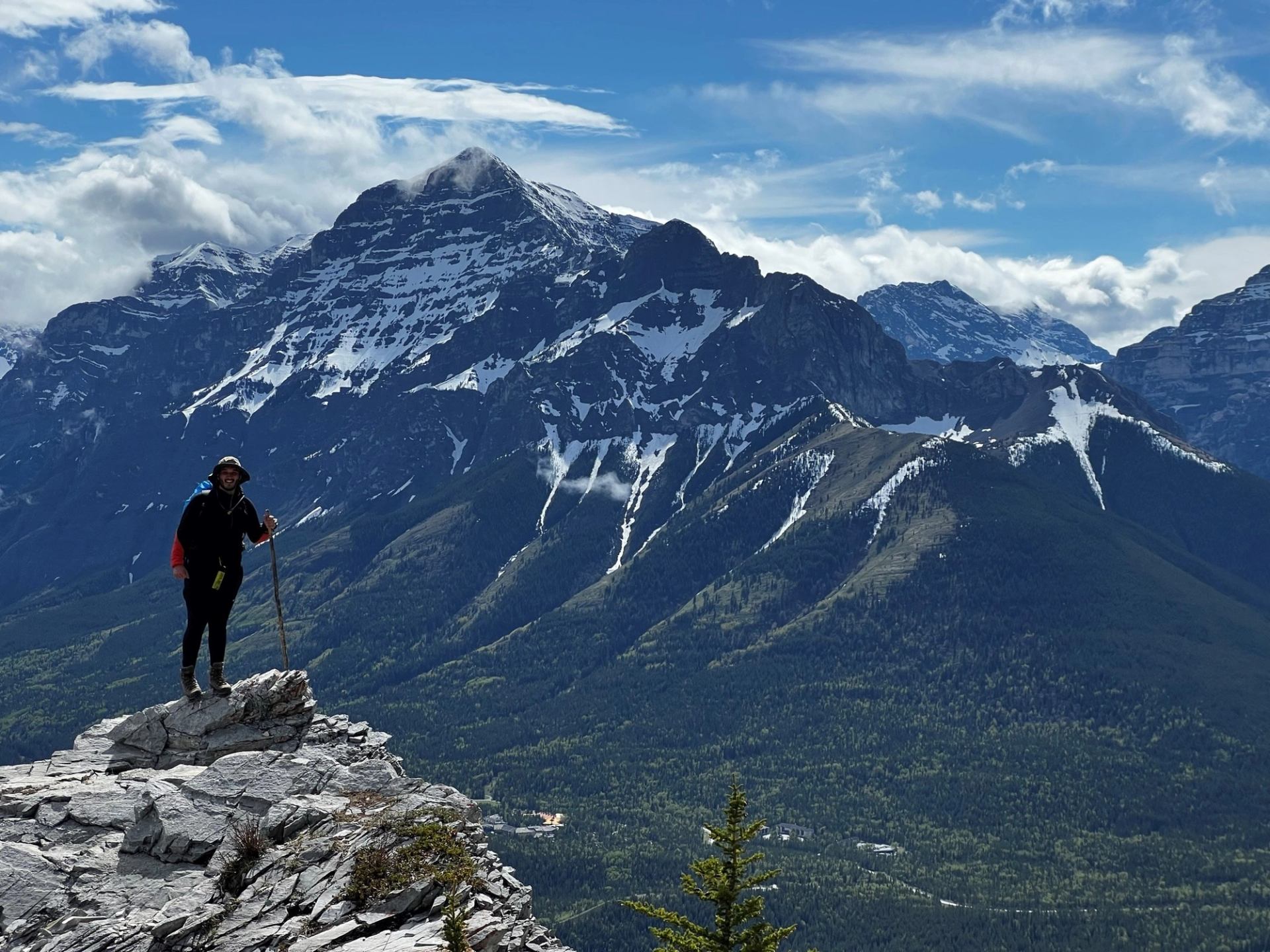 Hiker on rocky ledge overlooking snowy green mountains.