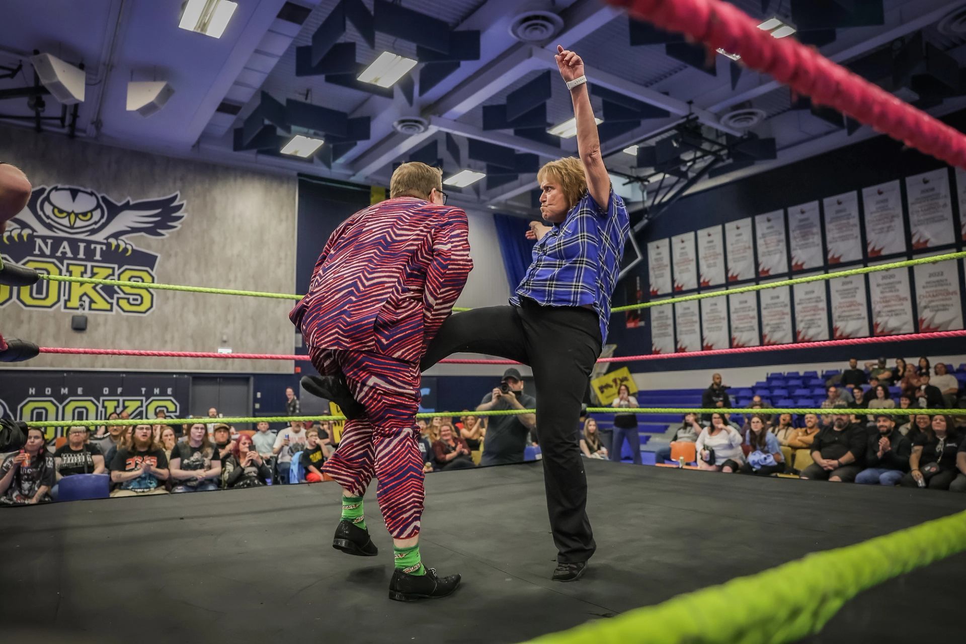 Wrestler in patterned suit takes kick from opponent in plaid shirt inside NAIT ring.