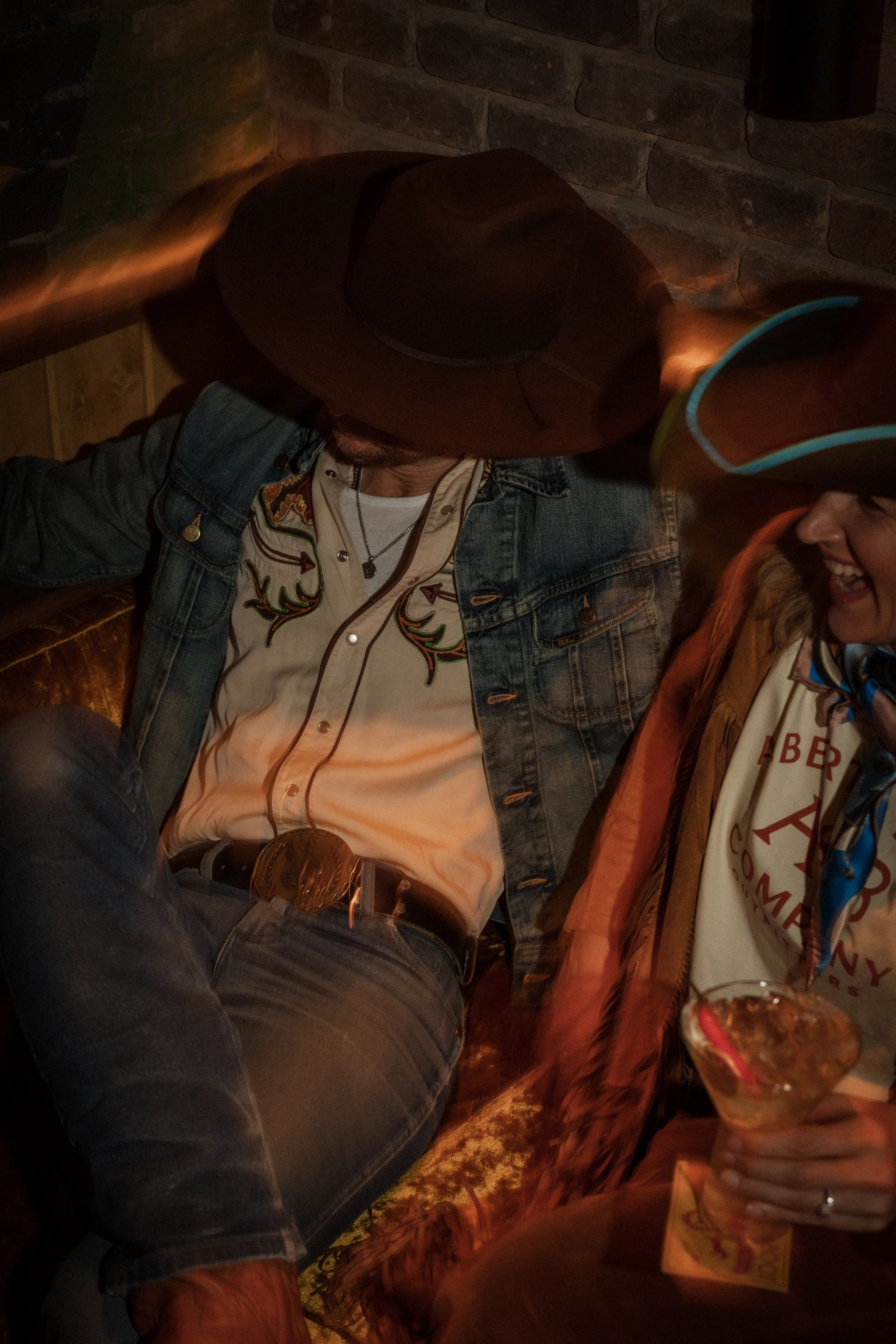 Two people relaxing on a couch, one in a denim jacket and cowboy hat, holding a drink.