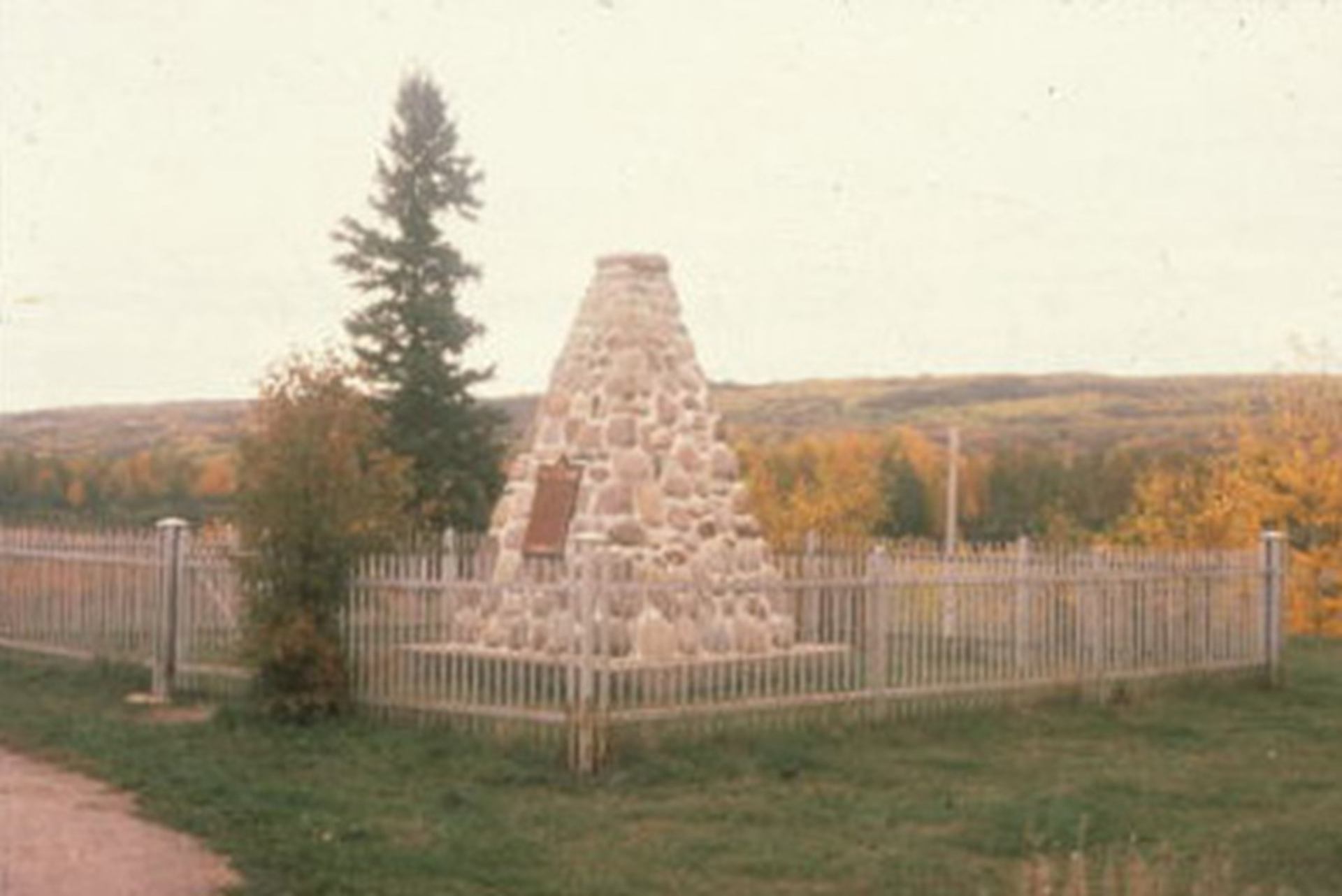 Stone cairn monument with plaque inside fenced area, trees and rolling hills behind.