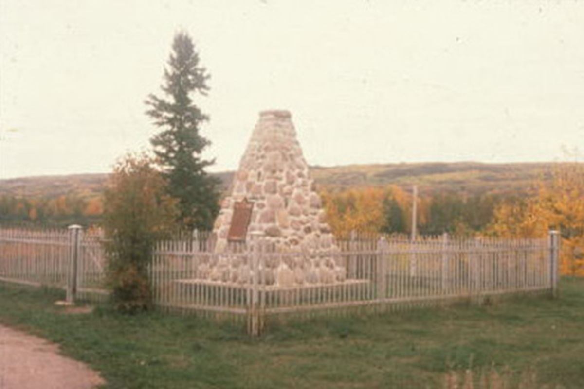 Stone cairn monument with plaque inside fenced area, trees and rolling hills behind.