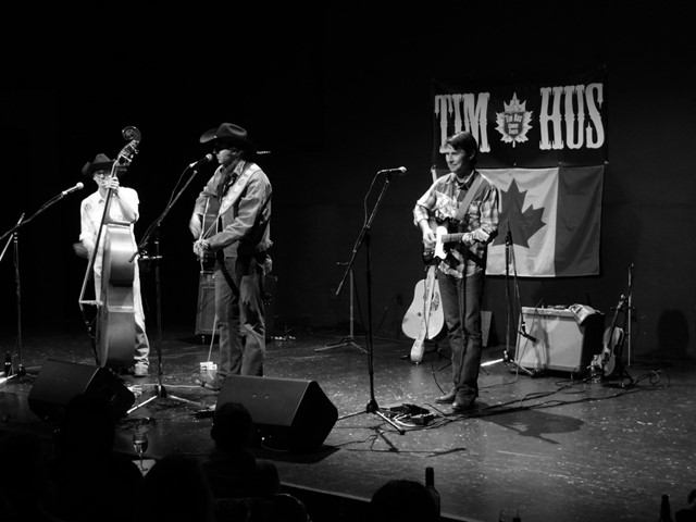 Country band performing on stage with guitars, bass, and microphones beside a flag backdrop.