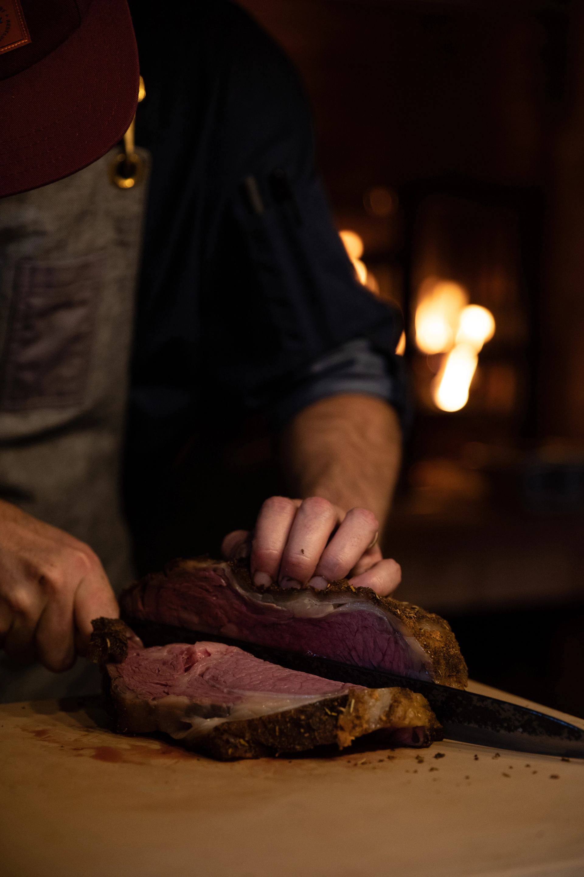 A person carves a pink-centered roast on a wooden board, with blurred warm lights in the dark background.