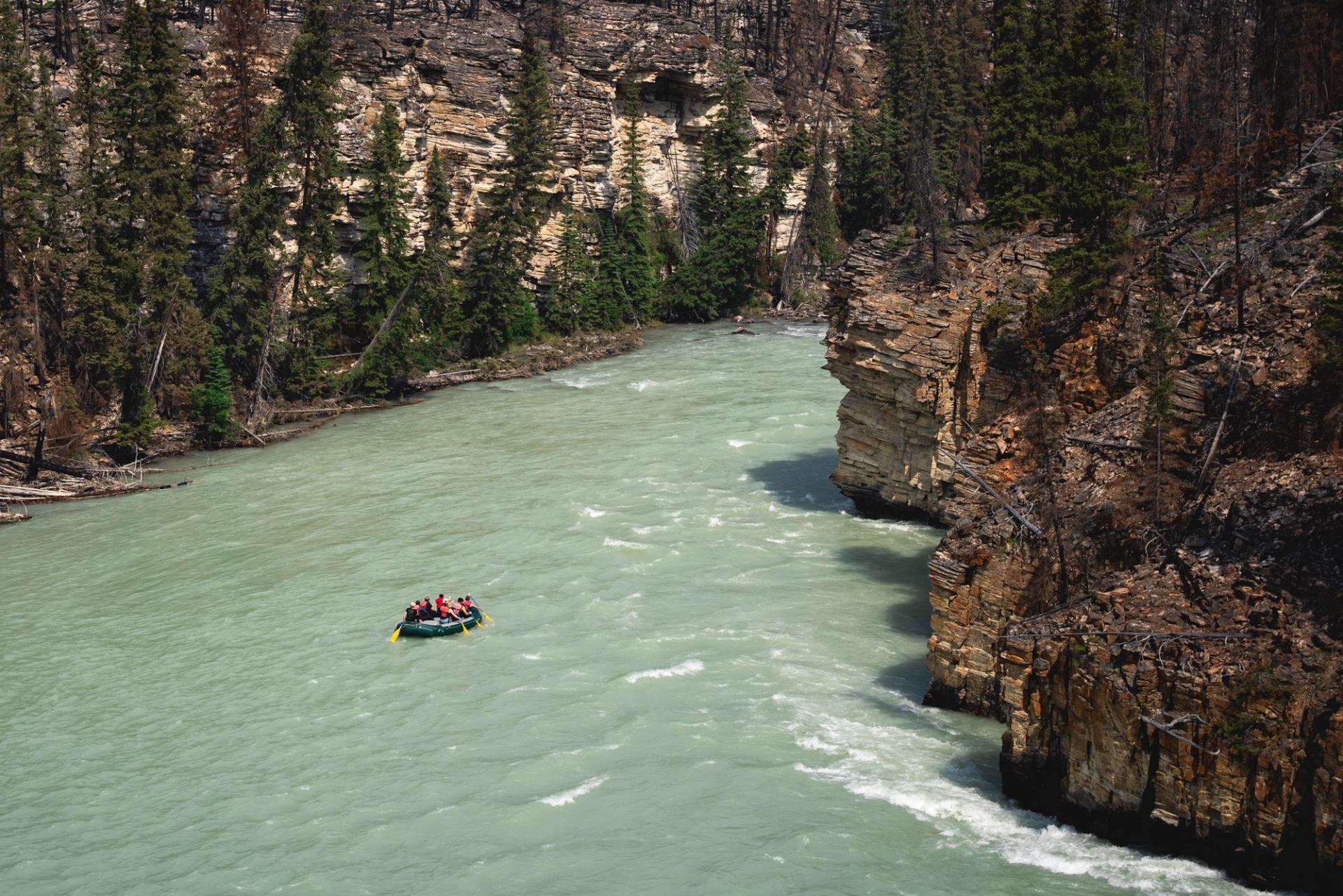 Raft floating down a wide river between tall canyon walls and evergreen trees.