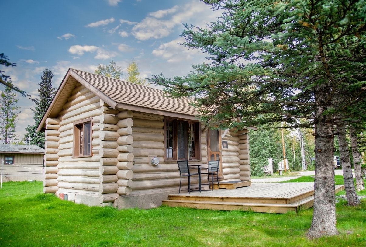 A rustic log cabin with a wooden deck sits on a grassy campsite surrounded by mature evergreen trees.