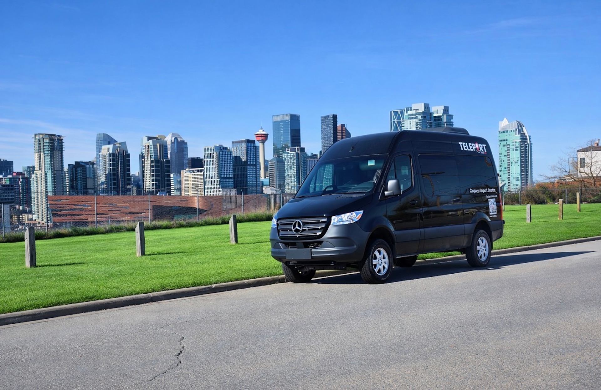 Black Teleport shuttle van parked by grass with Calgary skyline and tower behind