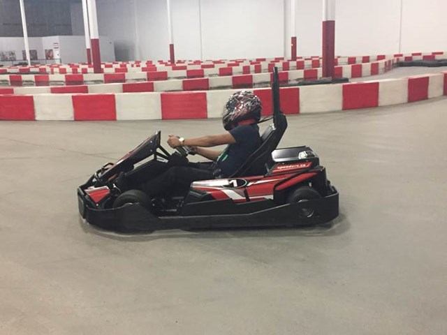 Racer in helmet driving go-kart on indoor track with red and white barriers.