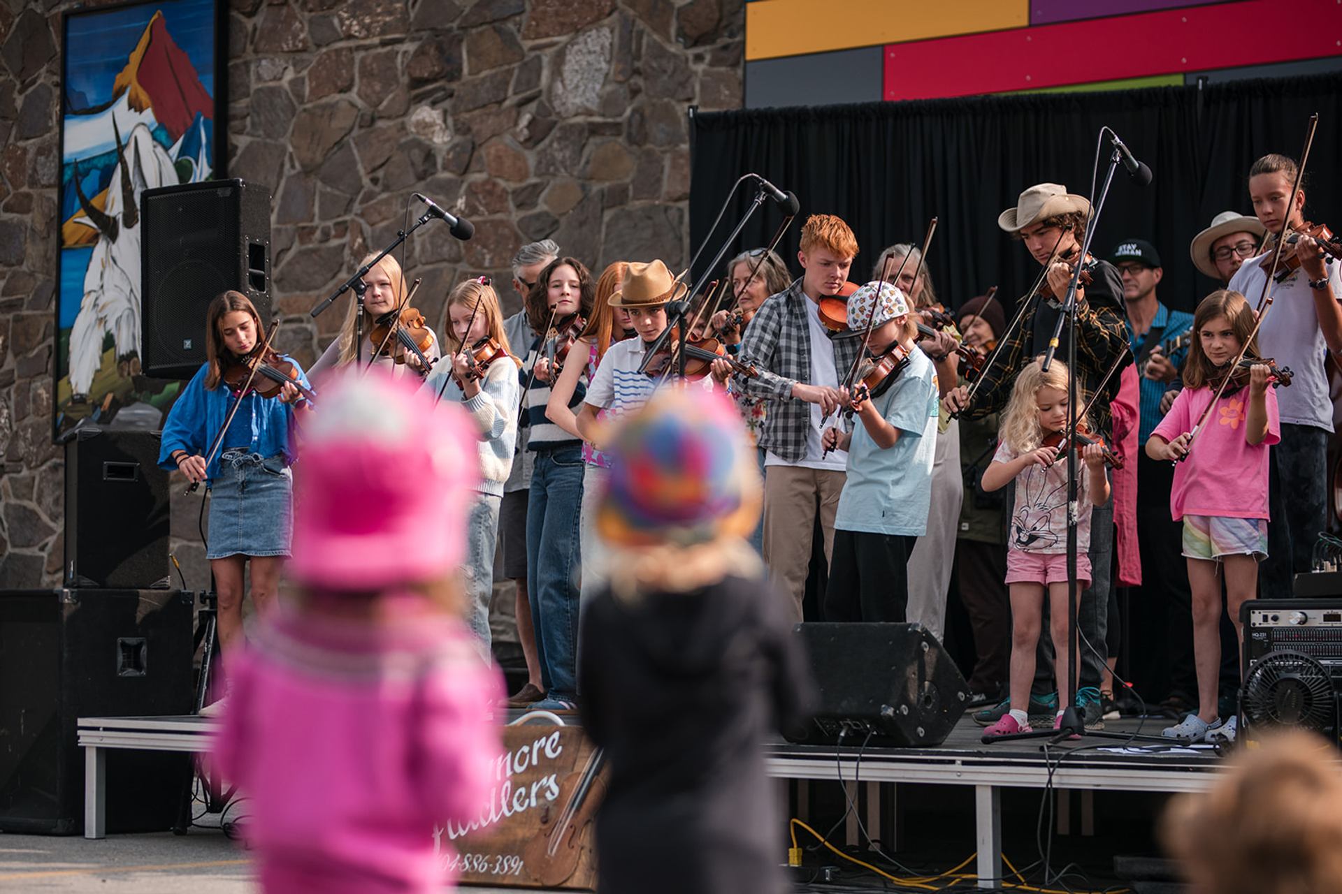 Two children watch as a group of fiddle players perform on stage.