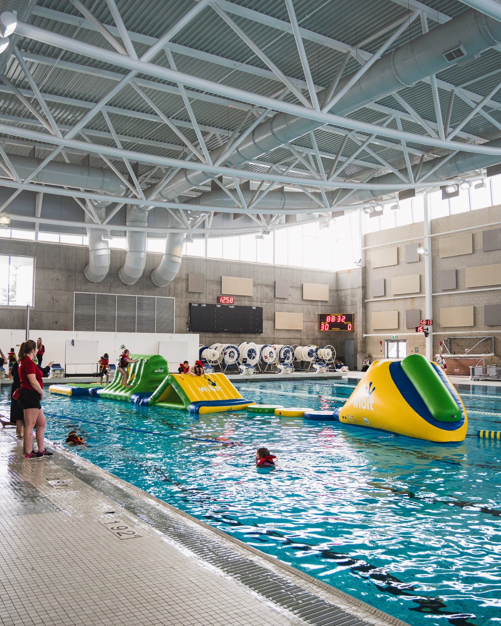 Indoor swimming pool with inflatable play structures, spectators, and scoreboard under steel roof tr.