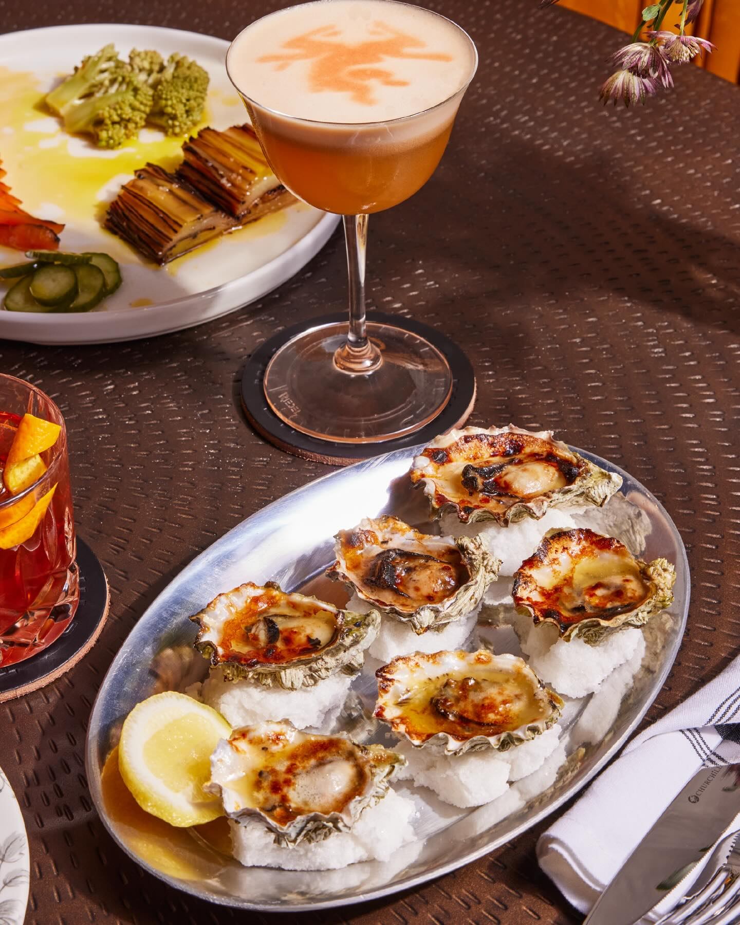 A platter of baked oysters on ice, a cocktail with an orange dragon design on foam, and other dishes on a table.