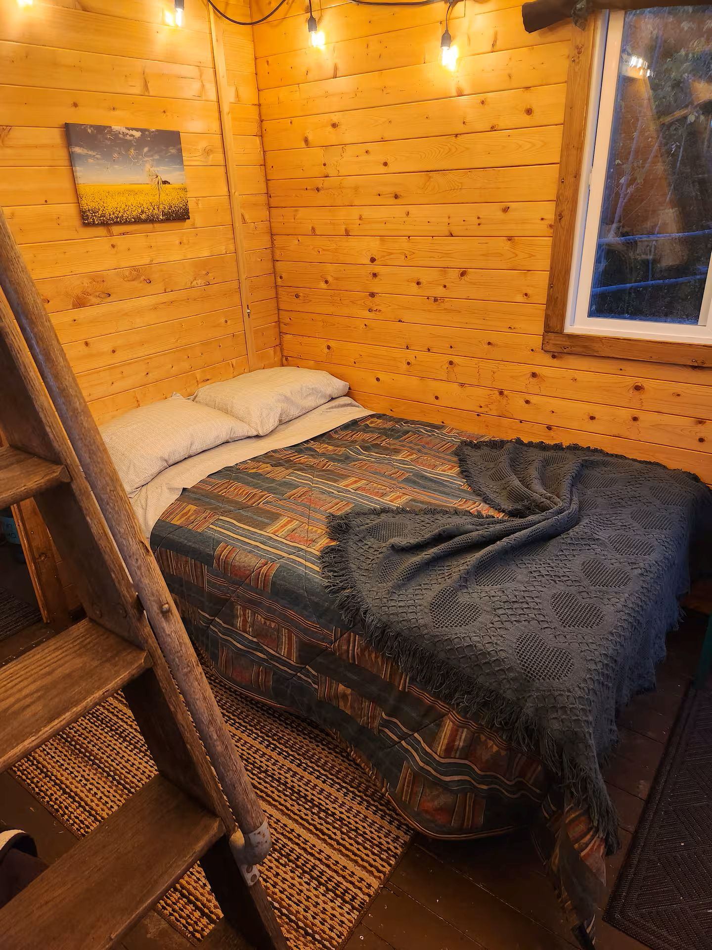Bed in a wood‑paneled cabin corner with string lights and a window.