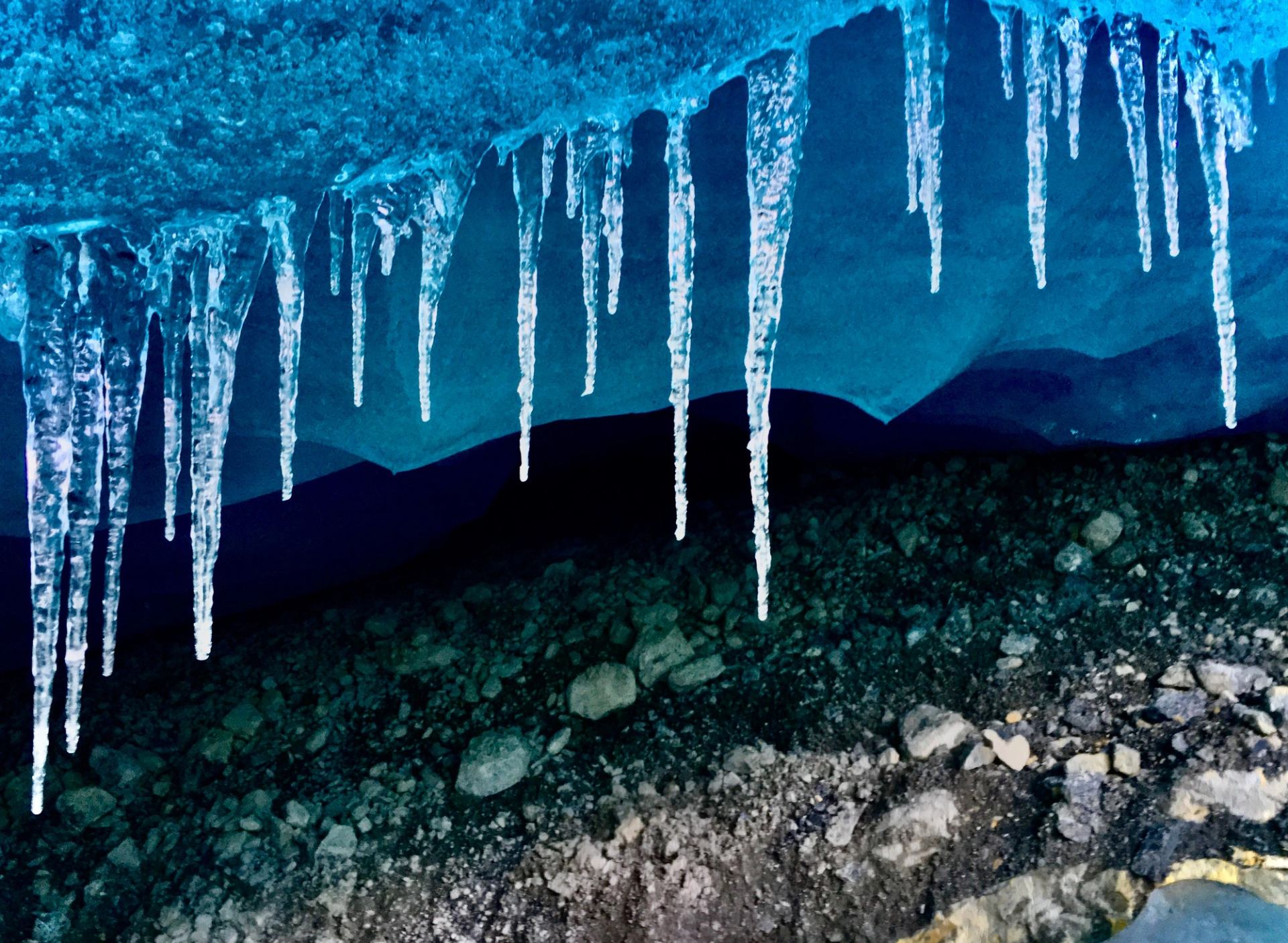 Blue ice cave ceiling with hanging icicles above rocky ground.