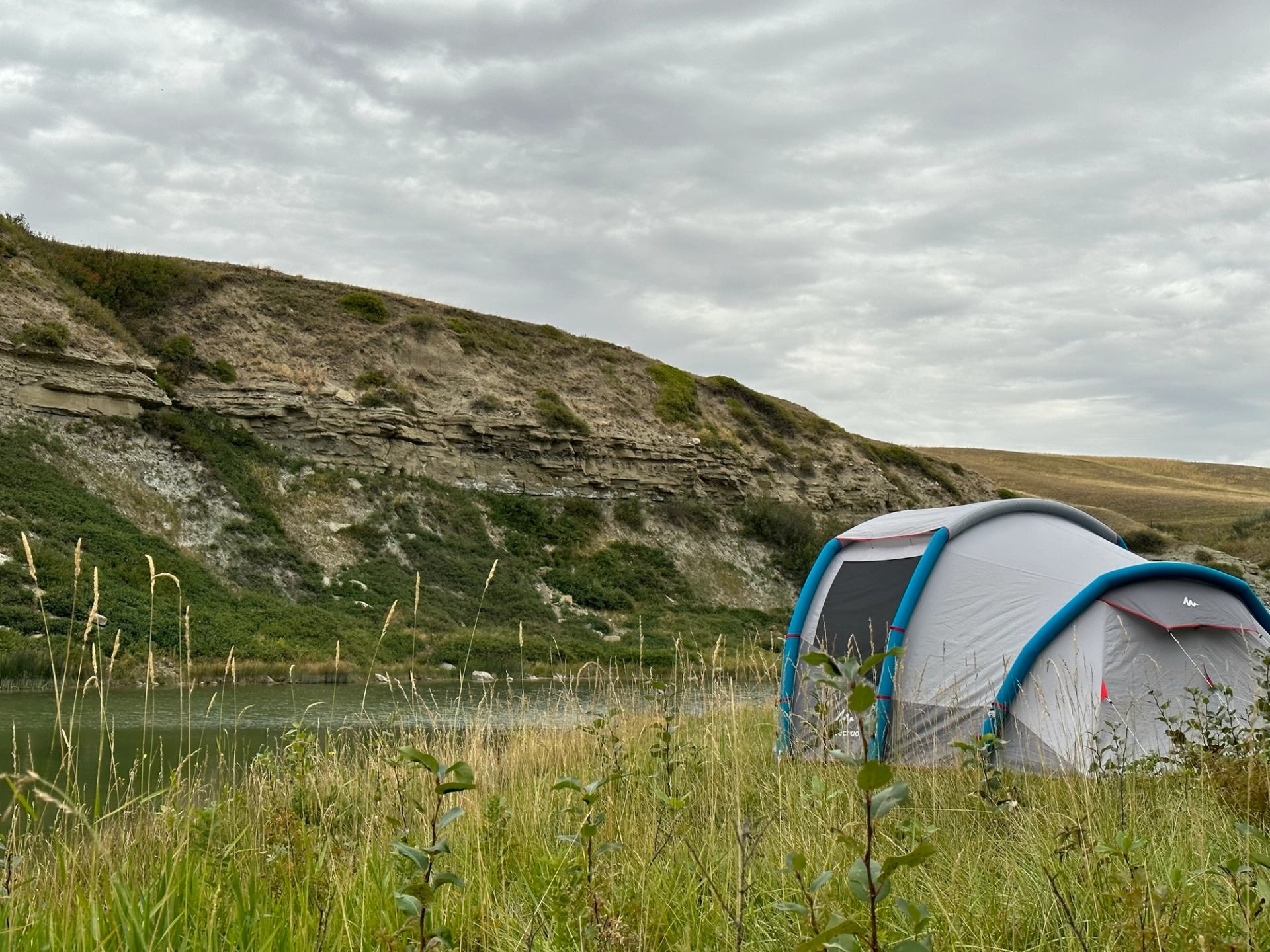 A tent set up beside a calm river with cliffs and grassy hills behind it.