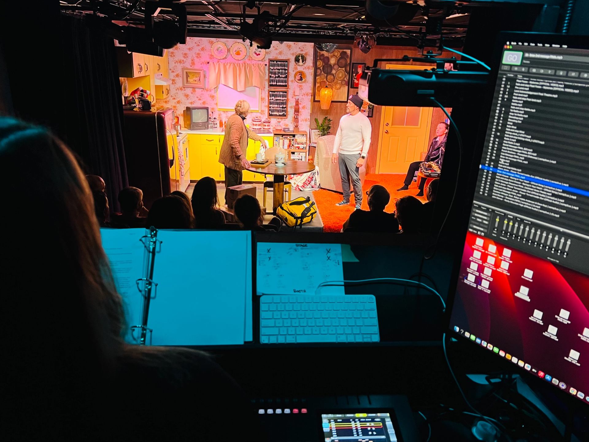 Tech crew monitors stage as actors perform amid colorful props.