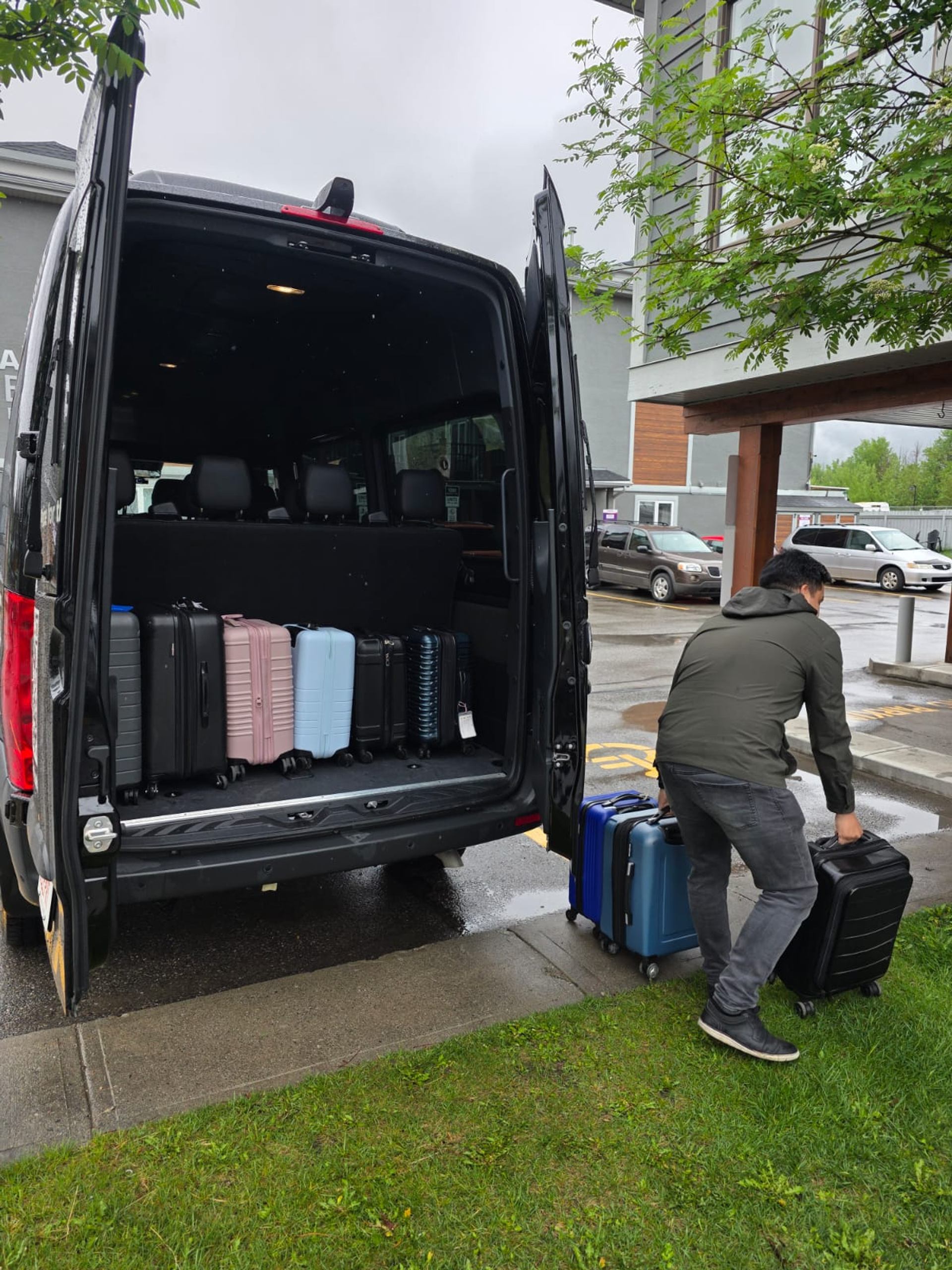 Person loads rolling suitcases into open black van outside a building entrance.