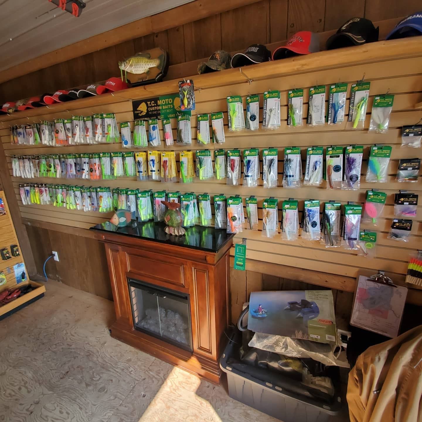 Fishing tackle display wall with packaged lures, hats above, and small electric fireplace below.