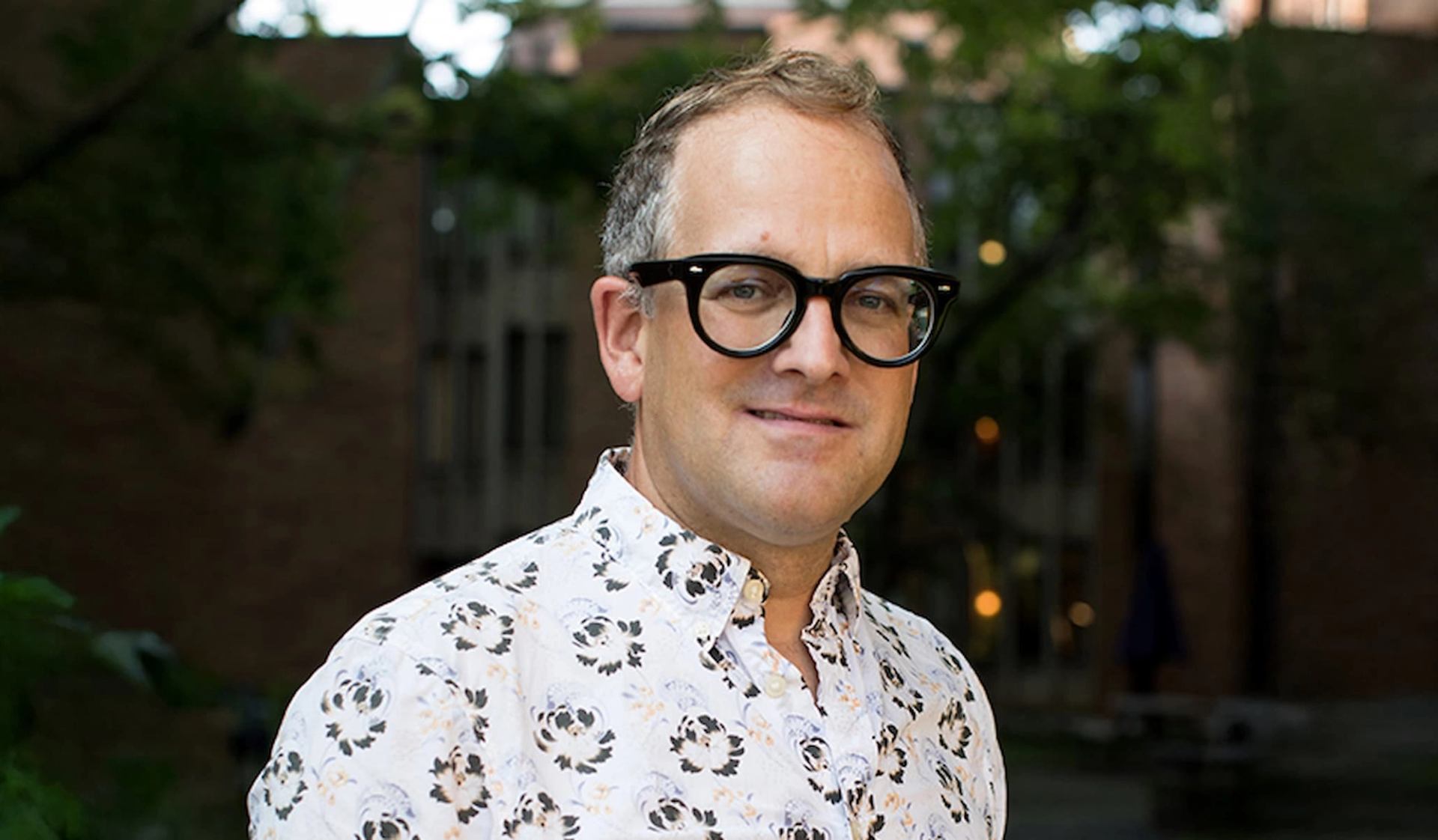Person wearing black glasses and floral shirt stands outdoors before blurred trees and buildings.