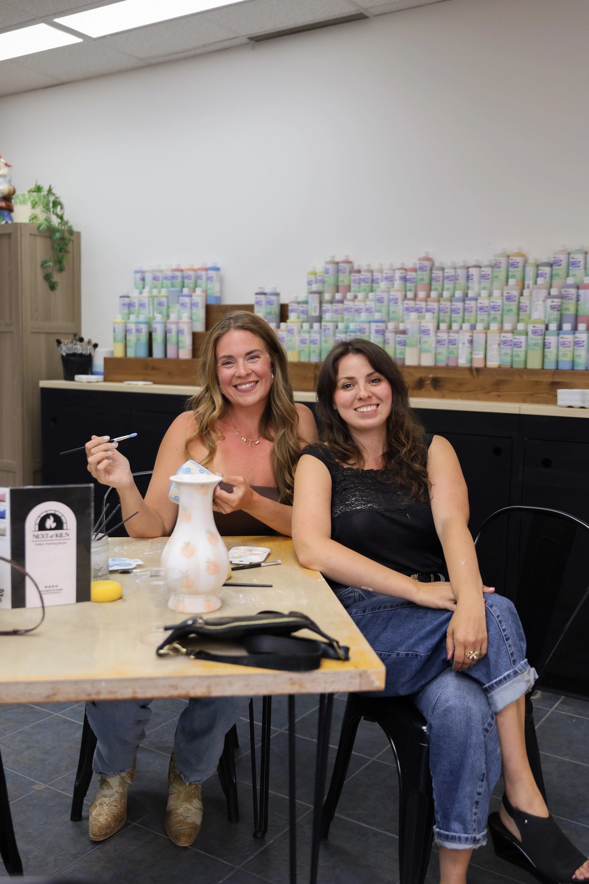 Two people sit at a pottery studio table, painting a ceramic vase together.