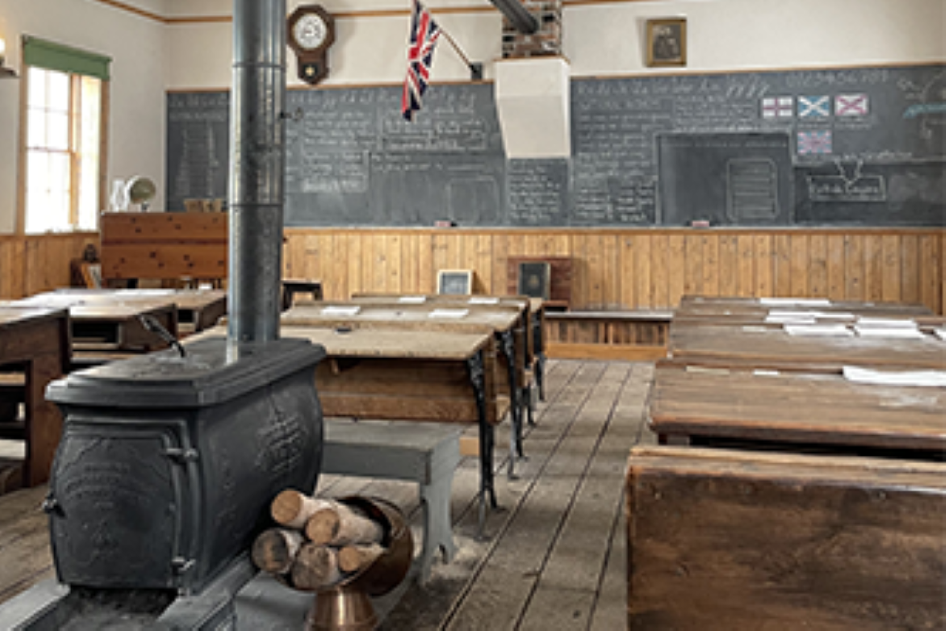 Historic classroom with wood stove, wooden desks, chalkboard notes and flag on wall.