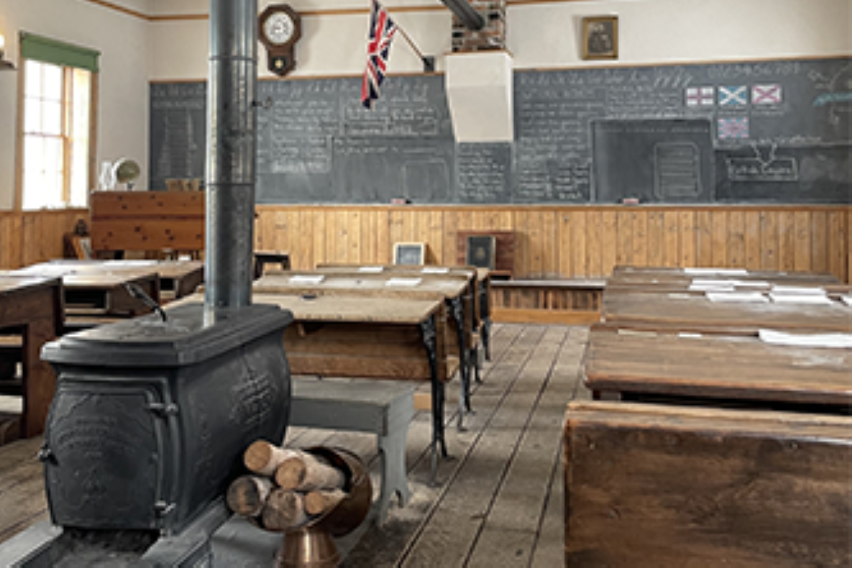 Historic classroom with wood stove, wooden desks, chalkboard notes and flag on wall.