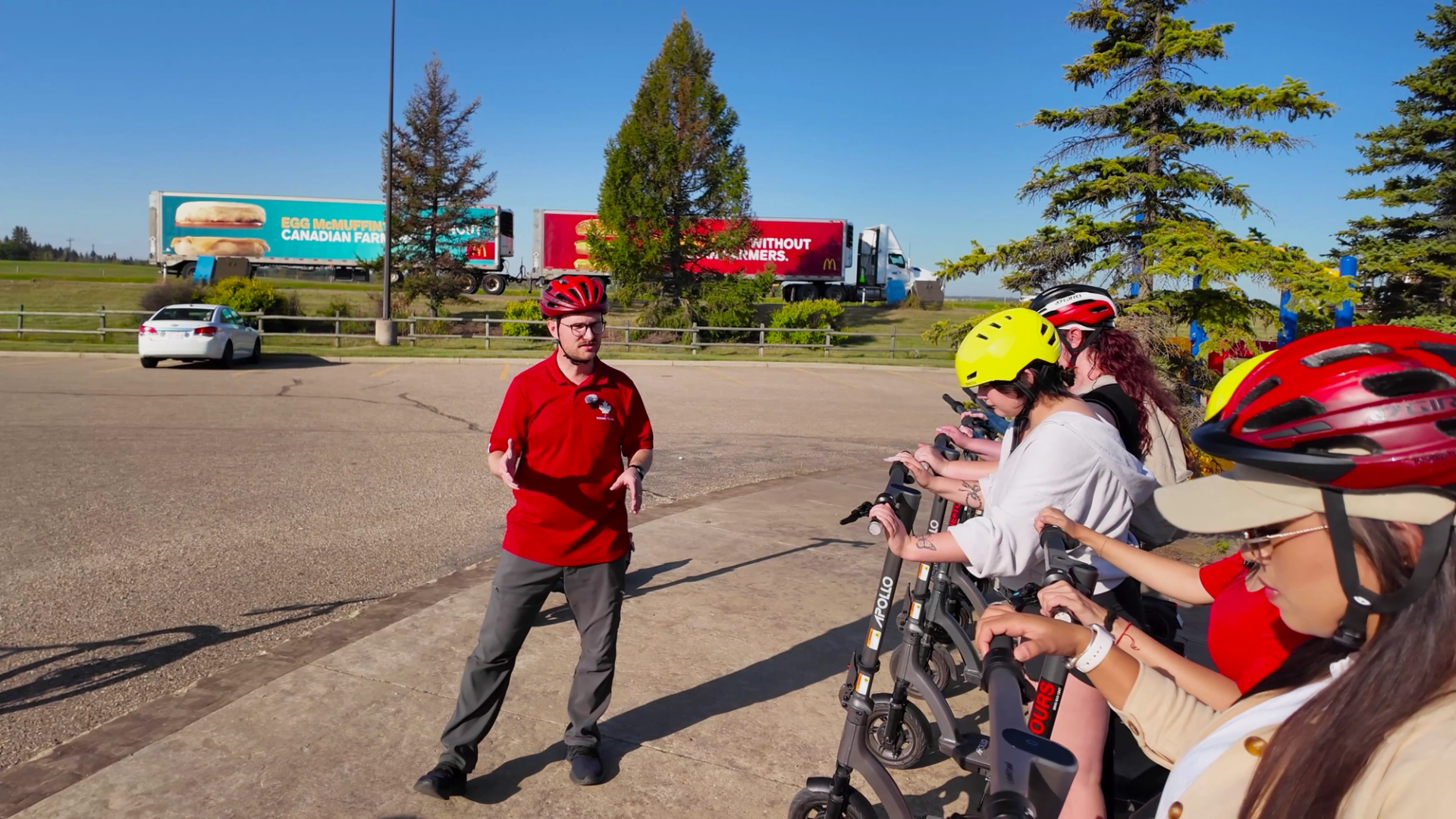 Instructor in red helmet talks to scooter riders wearing helmets in parking lot.