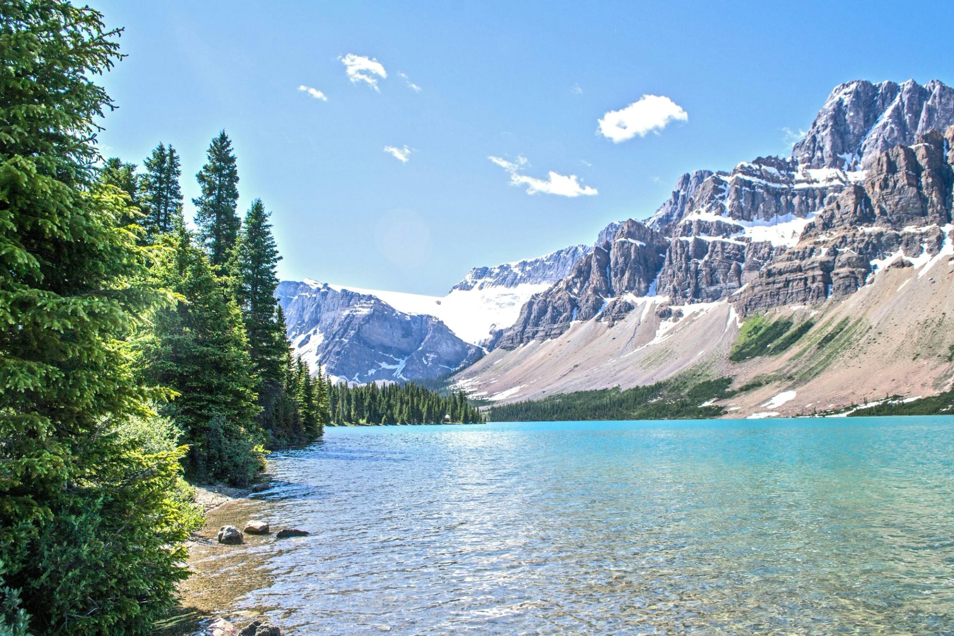 Turquoise mountain lake with pine trees along the shore and snow-capped peaks in the background.